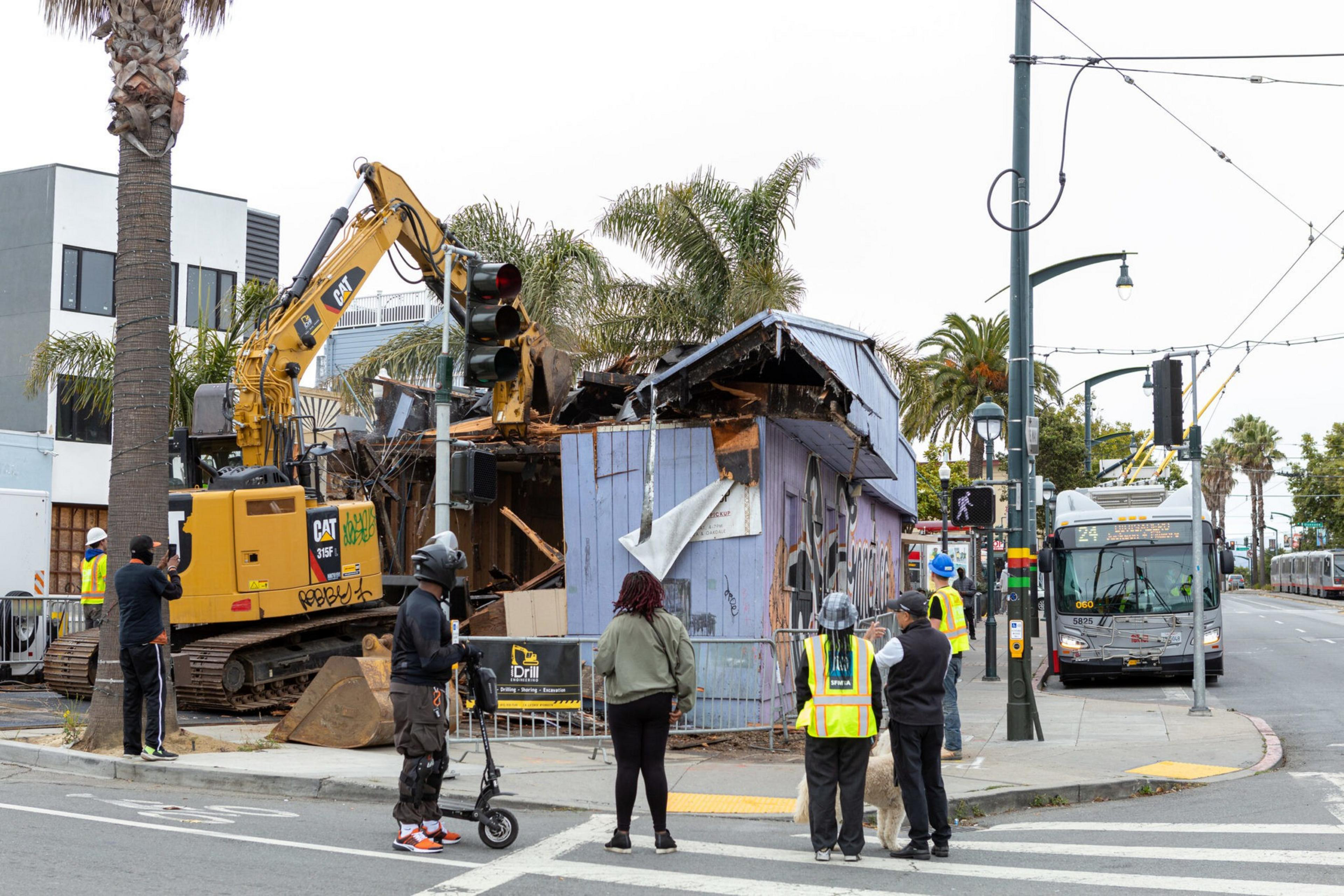 A small purple building is being demolished by a yellow excavator while people wait at the crosswalk, with a bus and palm trees in the background.