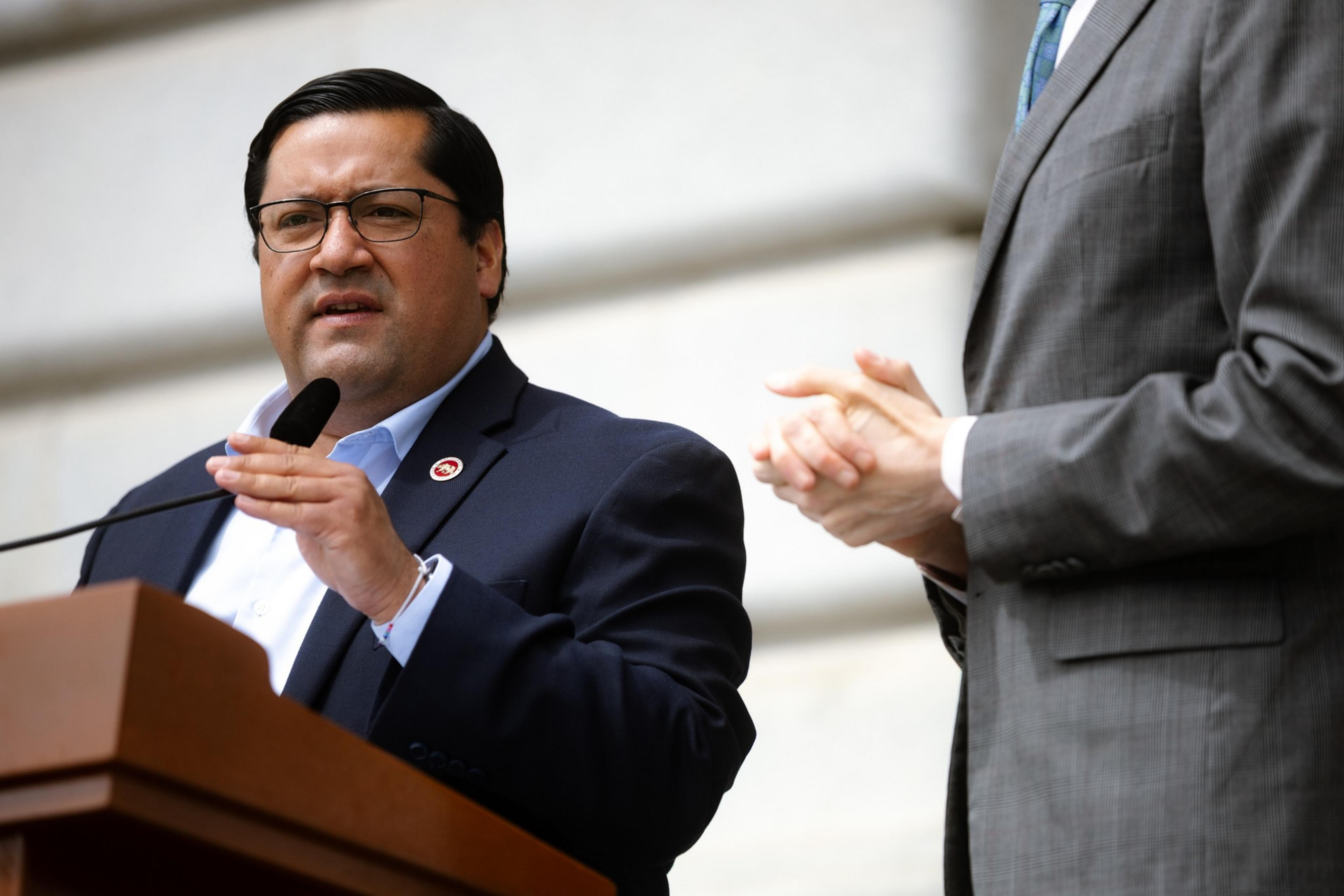 A man in a suit speaks at a podium with a microphone, while another person in a suit stands nearby with hands clasped. The background is a light, blurred structure.