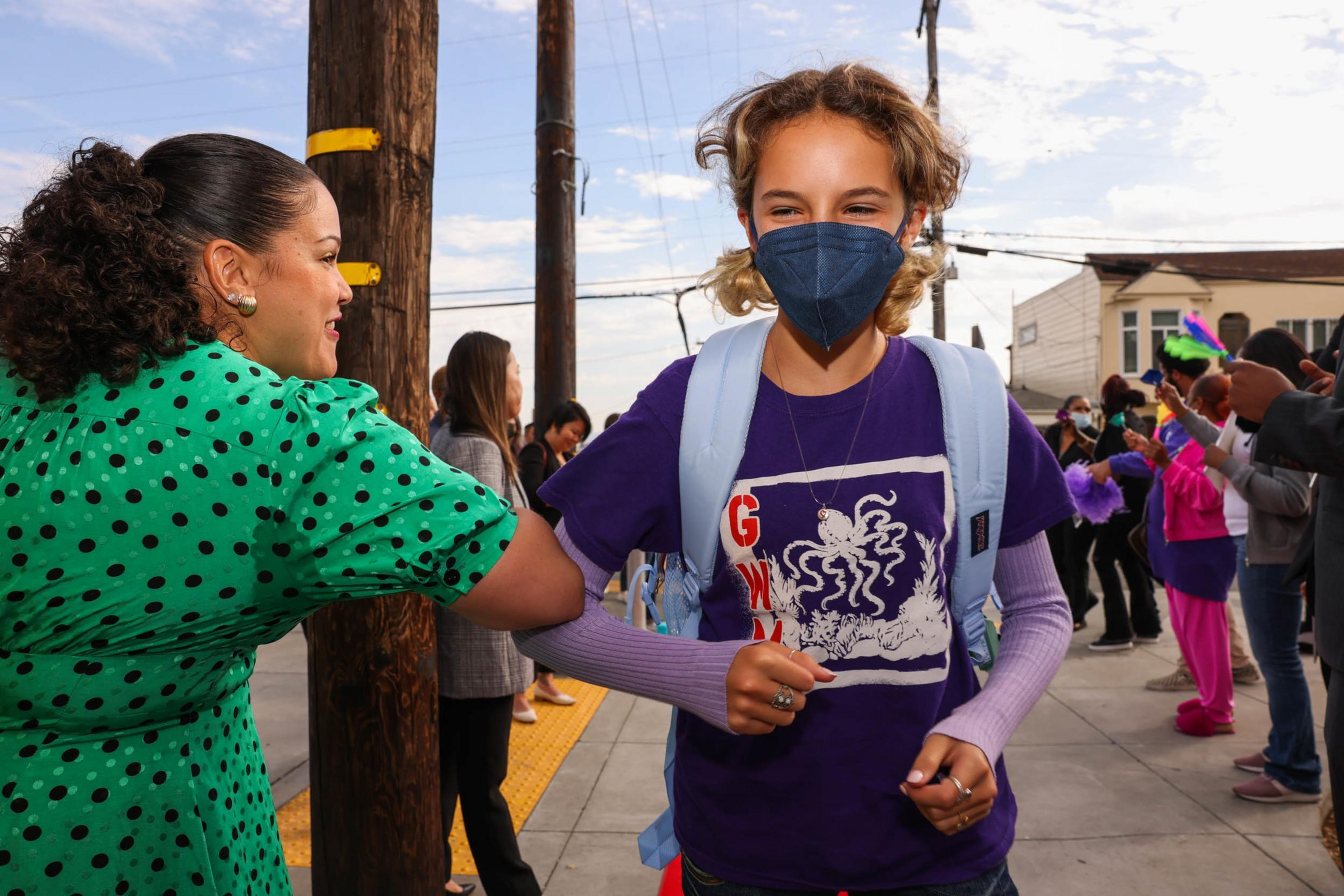 A girl with a blue face mask, purple shirt, and backpack bumps elbows with a woman in a green polka-dot dress on a busy sidewalk.