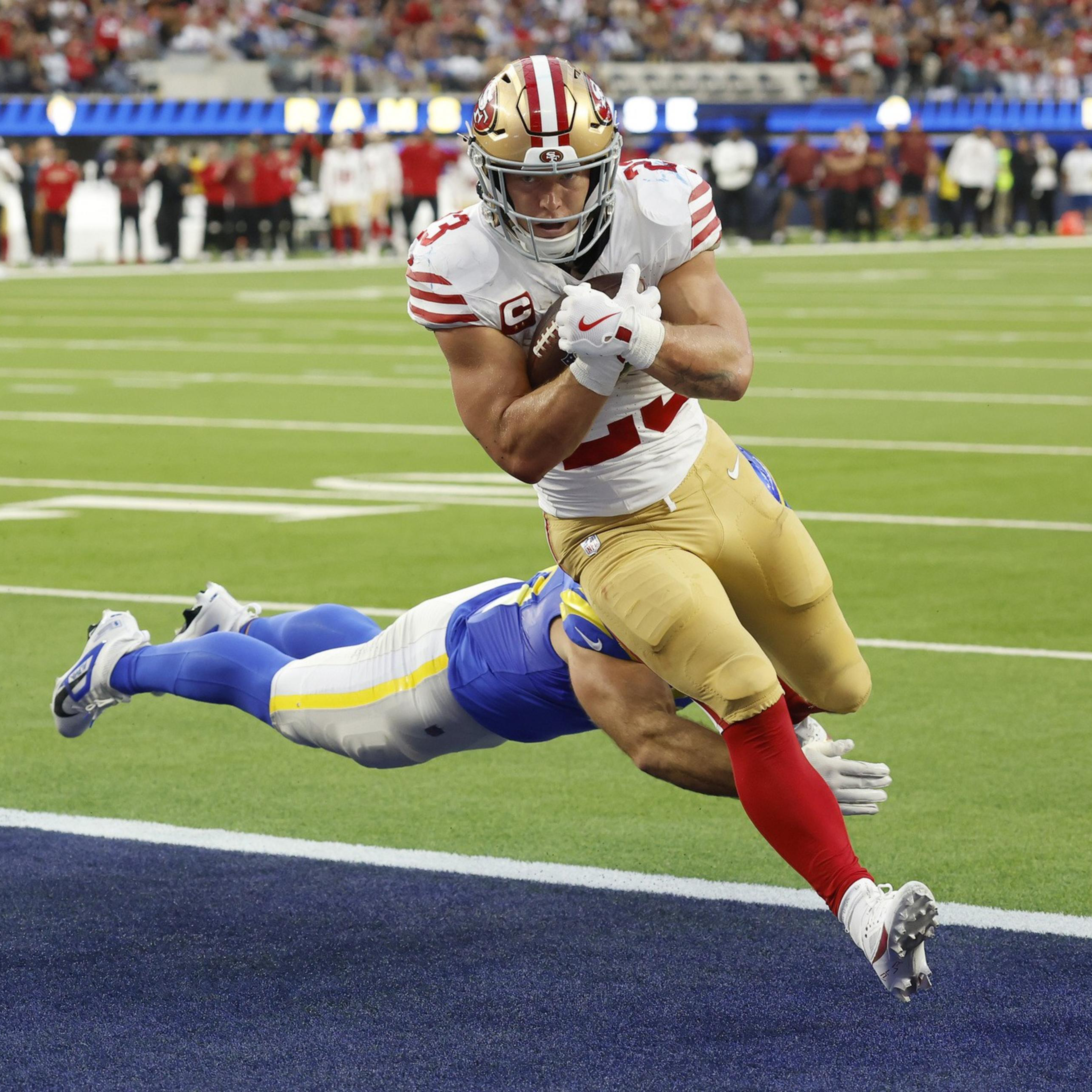 A football player in a white and red uniform leaps towards the end zone while a player in blue tries to tackle him mid-air.