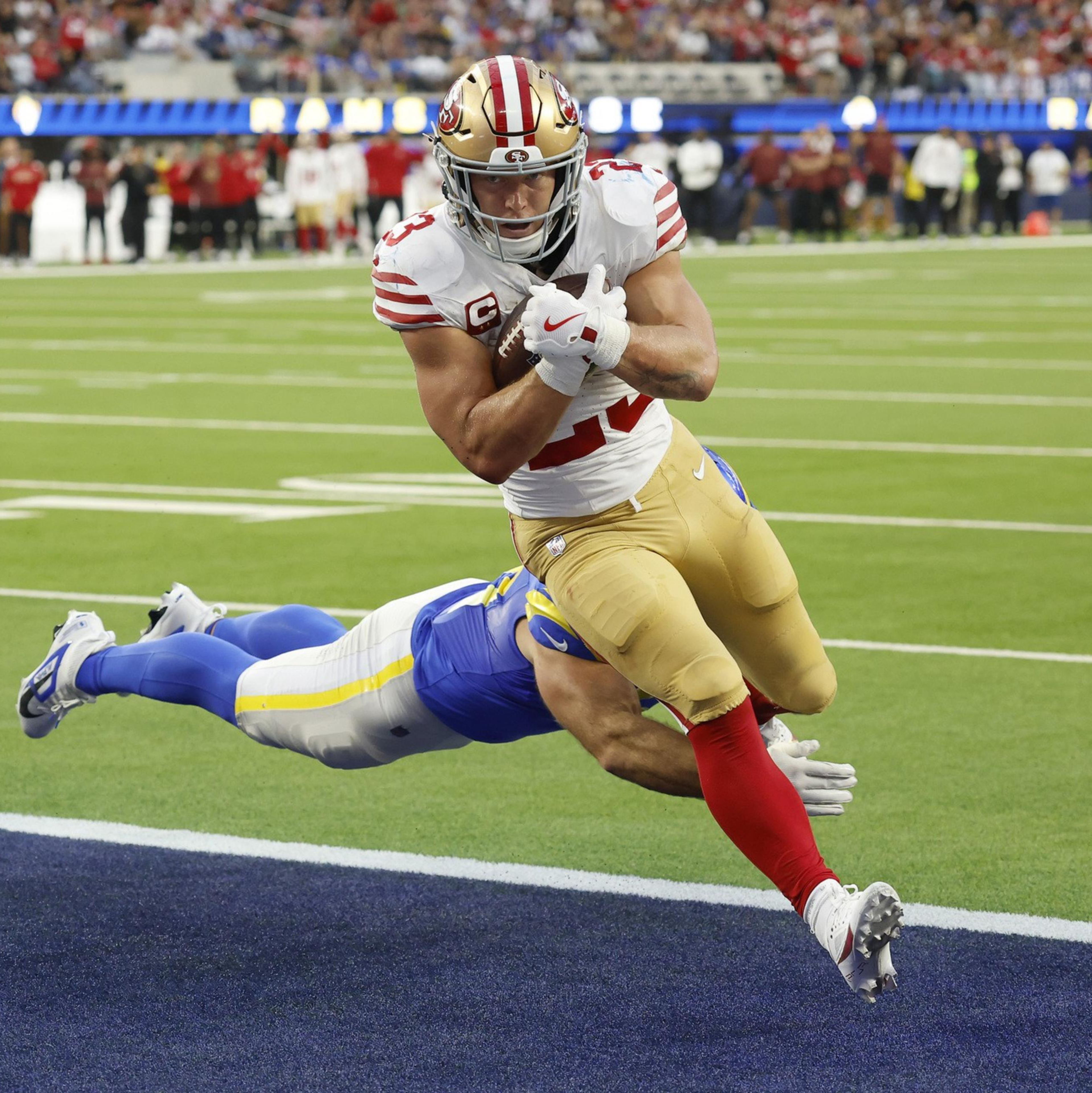 A football player in a white and red uniform leaps towards the end zone while a player in blue tries to tackle him mid-air.