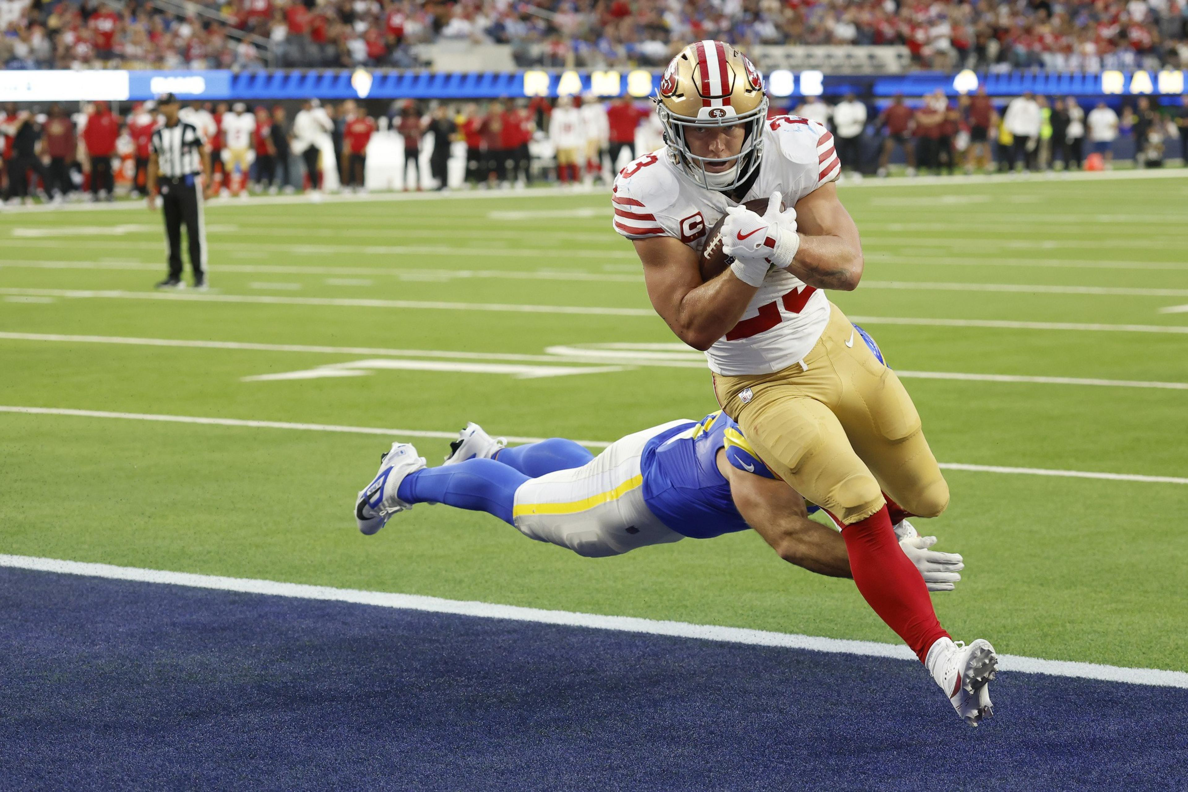 A football player in a white and red uniform leaps towards the end zone while a player in blue tries to tackle him mid-air.
