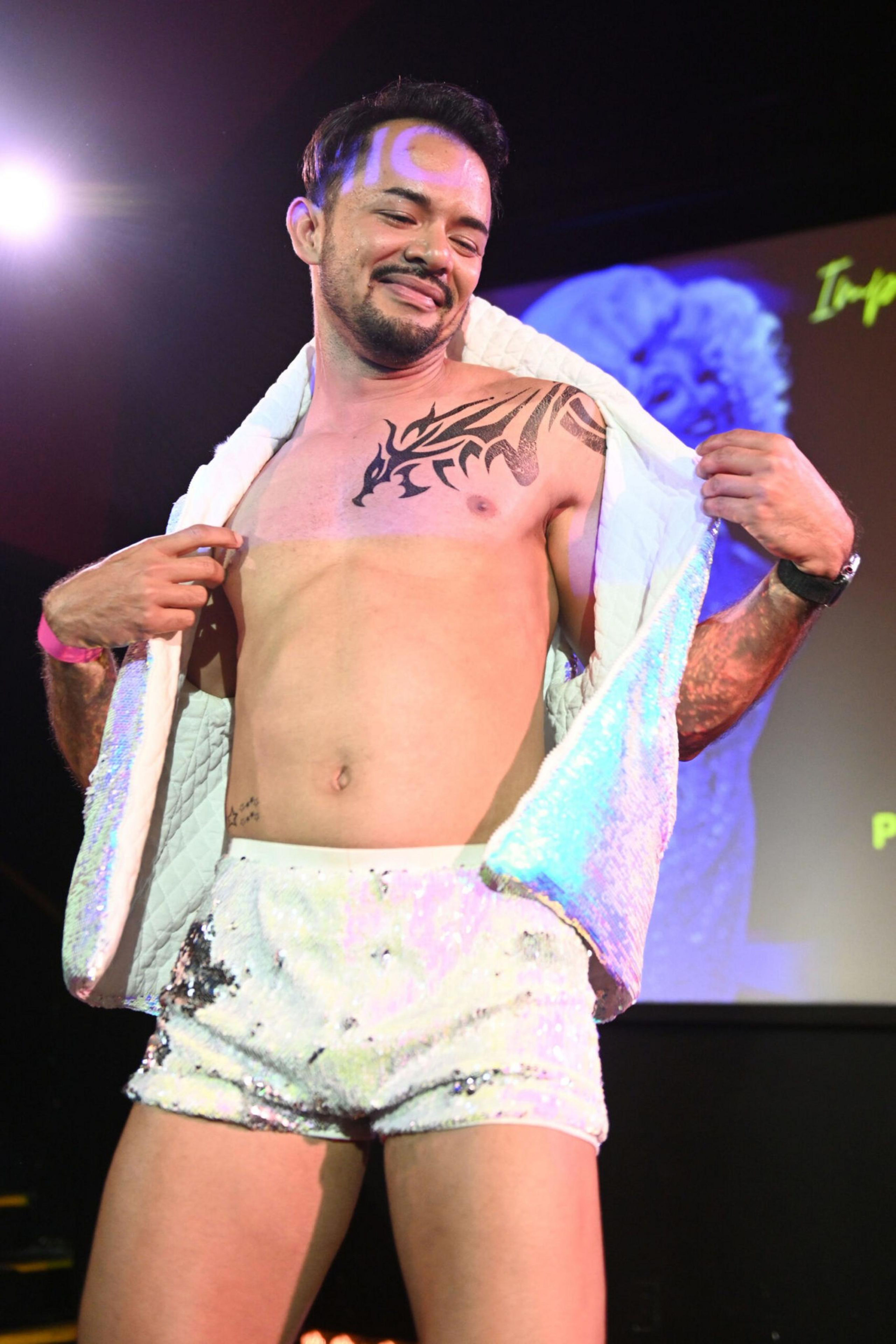 A man wearing white sequined shorts and a white open vest shows a tribal tattoo on his chest while smiling confidently under stage lights.