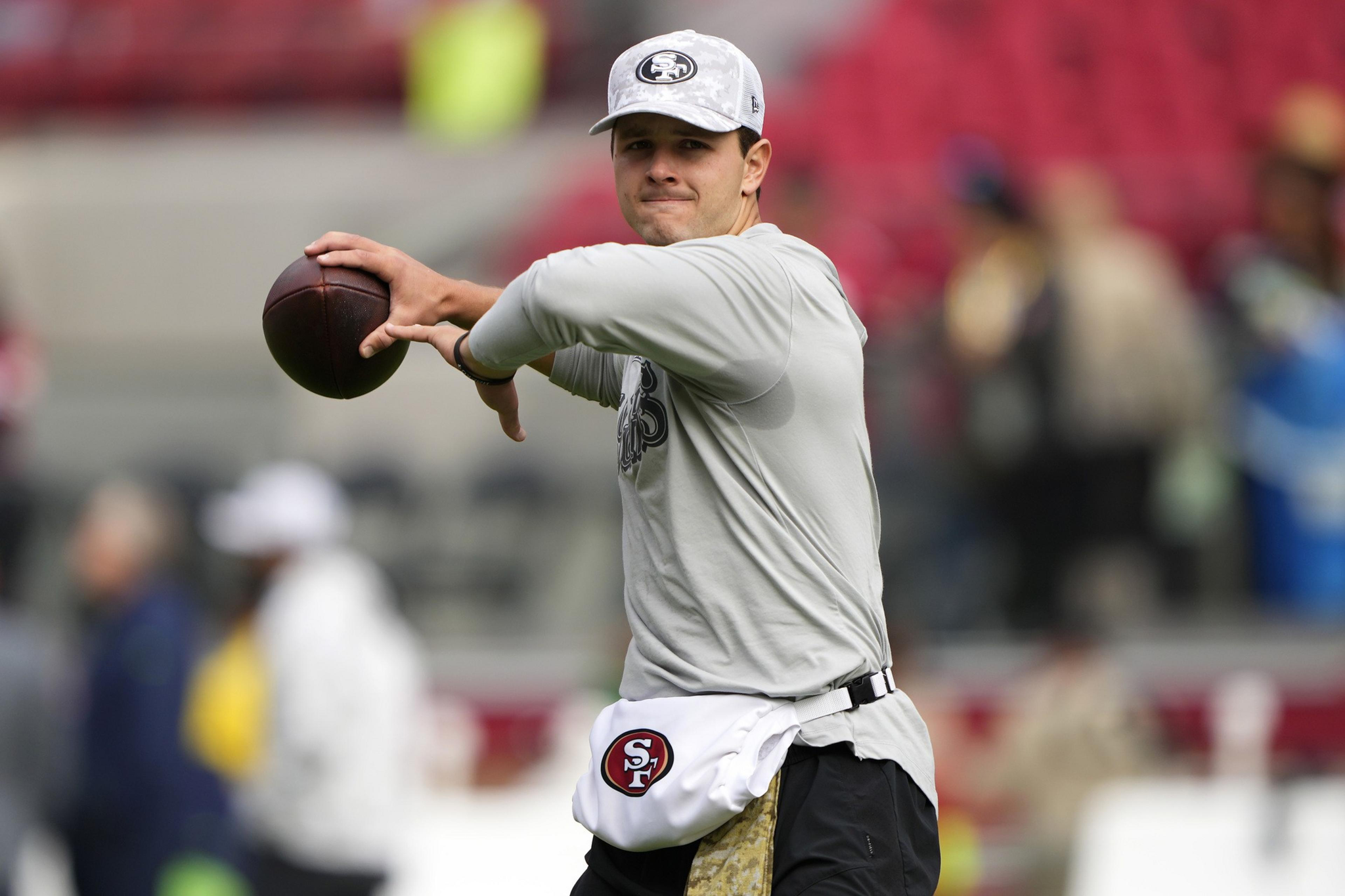 A person in a gray shirt and cap is preparing to throw a football. They wear a white pouch with a red and black logo. The background is a blurred stadium.