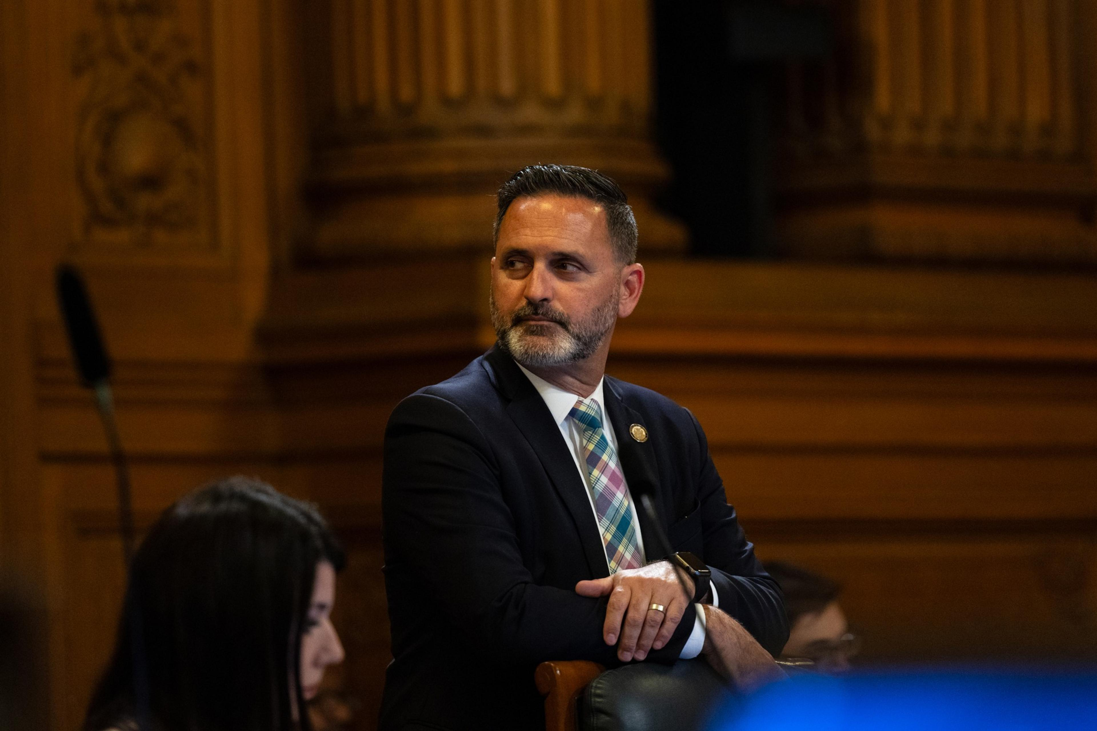 A man in a suit sits attentively, hands folded, in a chamber with ornate wood paneling.