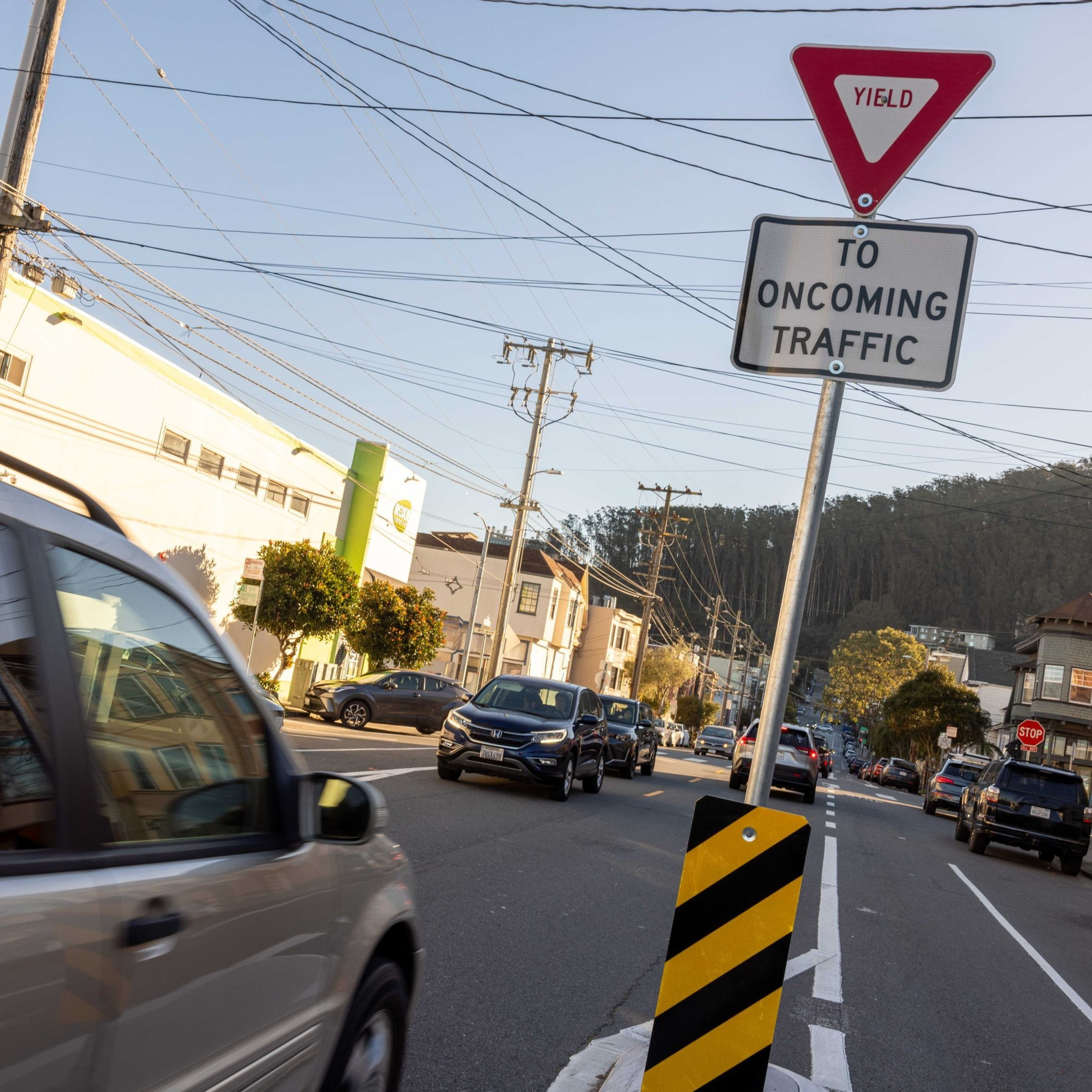 A street with cars passing by has a &quot;Yield to Oncoming Traffic&quot; sign beside a road divider, with buildings and trees in the background.