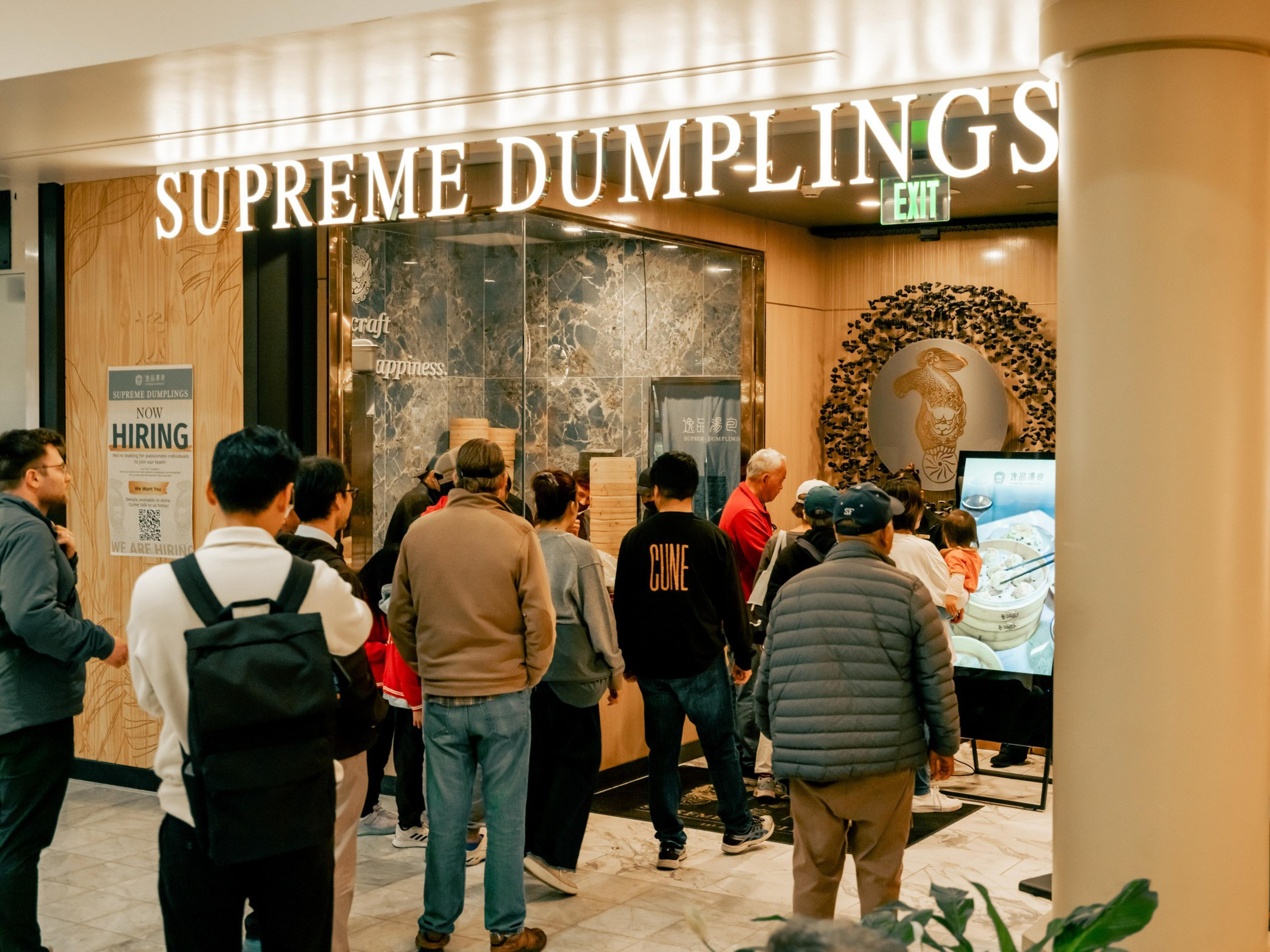 A group of people are lined up at a restaurant called &quot;Supreme Dumplings.&quot; The interior features marble decor and there's a sign indicating they're hiring.