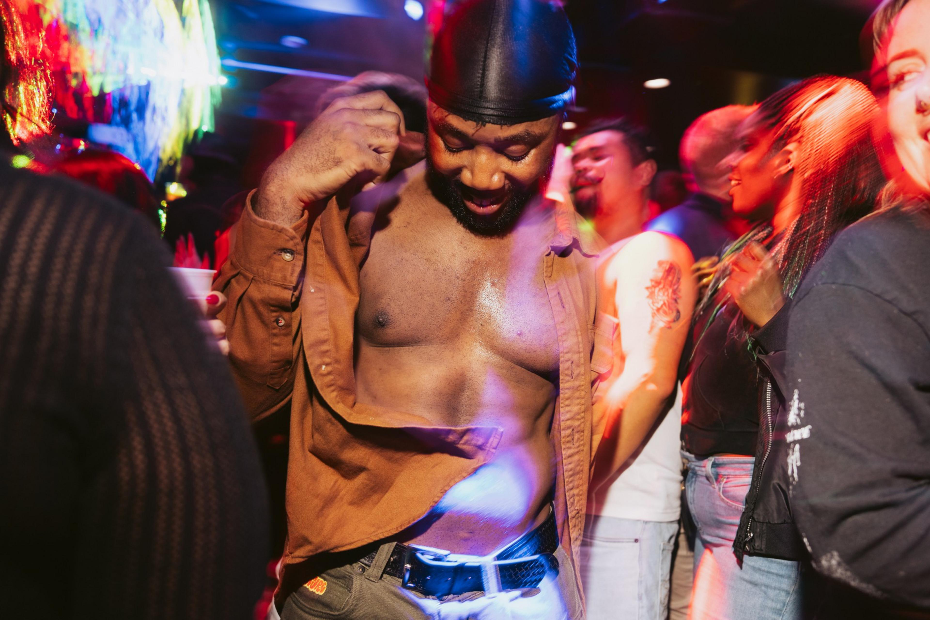 A man in a brown shirt, partially open, dances in a lively club filled with colorful lights and people. The atmosphere is vibrant and energetic.