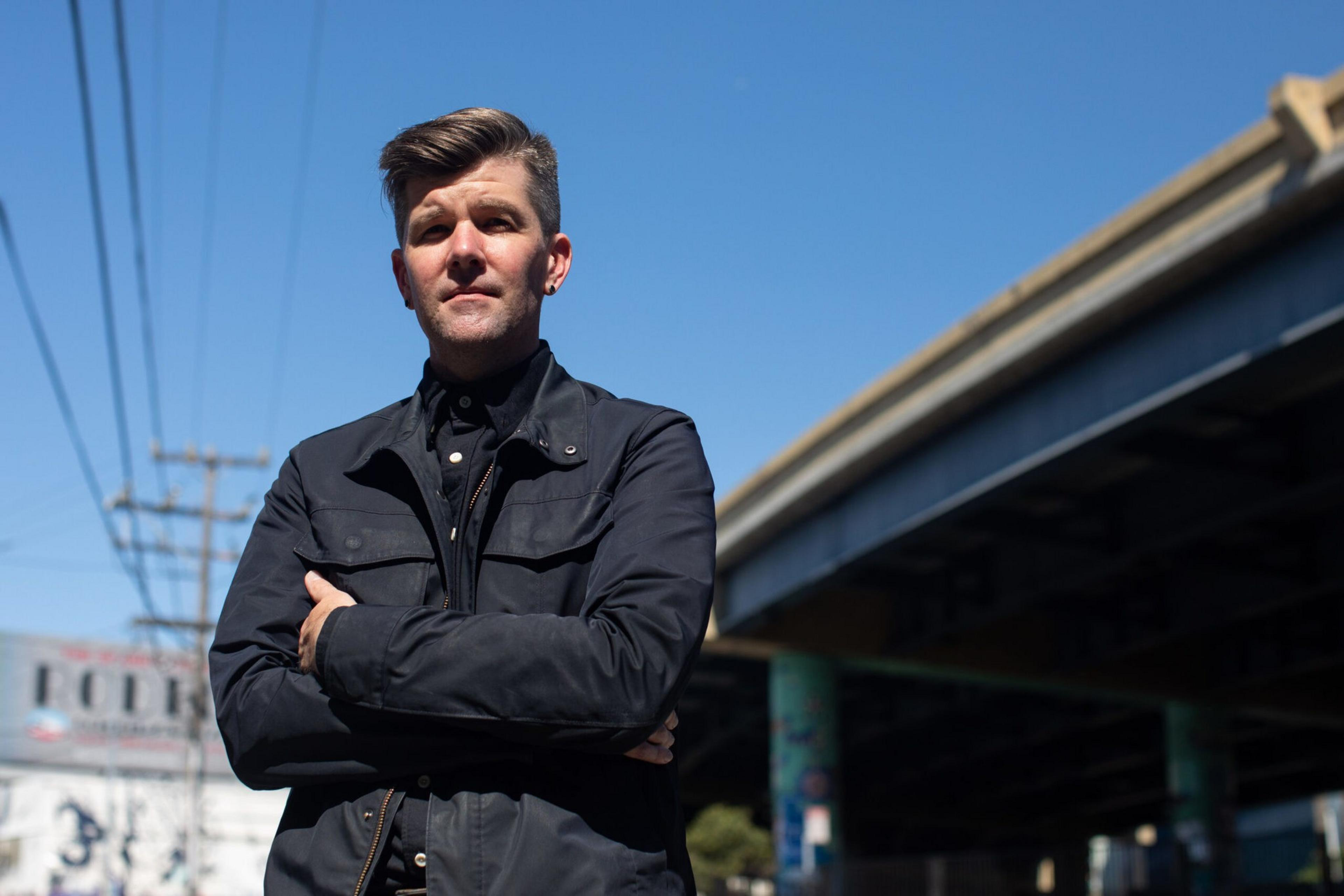 A man with light brown hair, wearing a dark jacket and shirt, stands with arms crossed under a clear blue sky near a bridge and power lines.