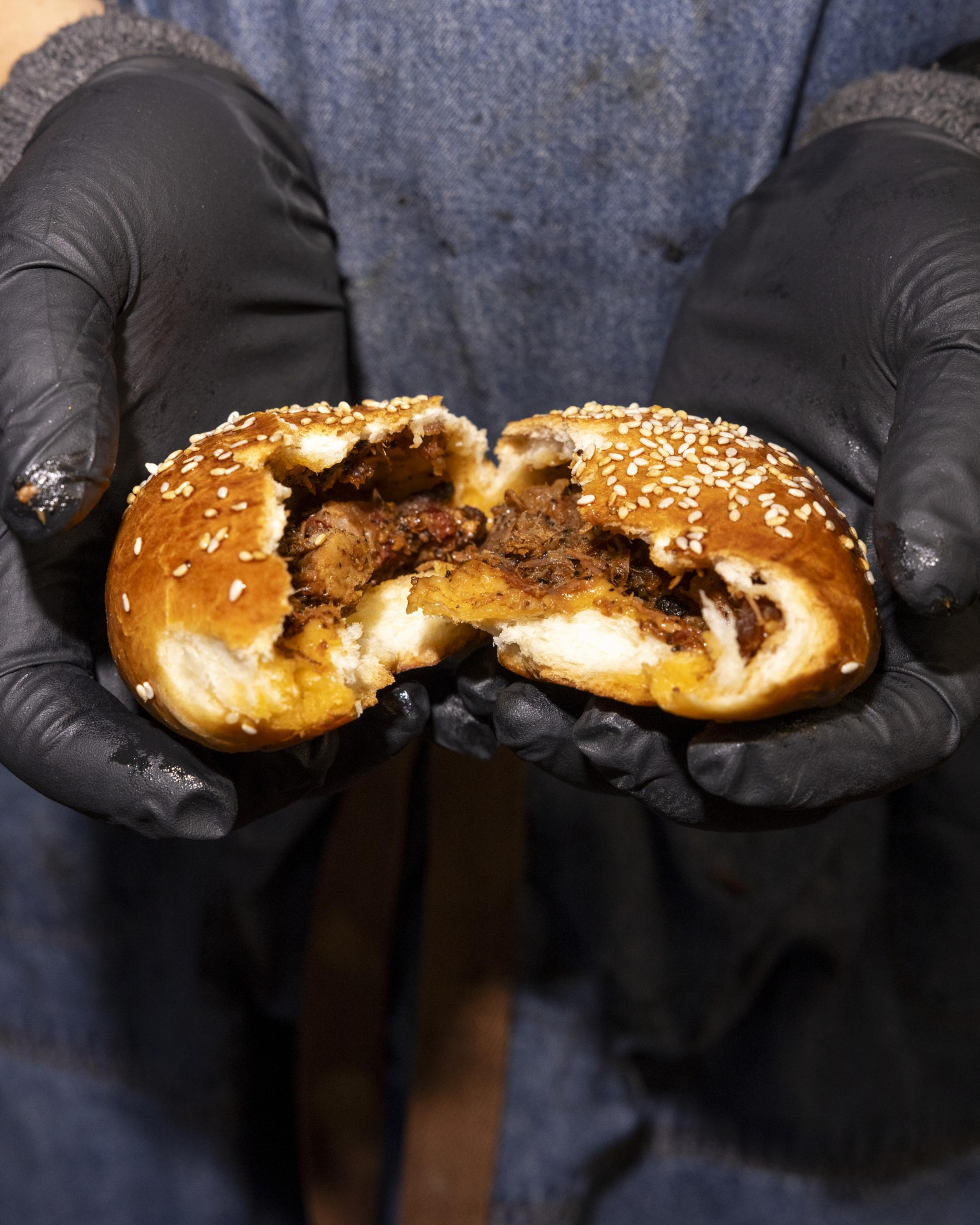 A person wearing black gloves is holding a sesame seed bun filled with pulled meat and melted cheese, torn open to show the tasty contents.