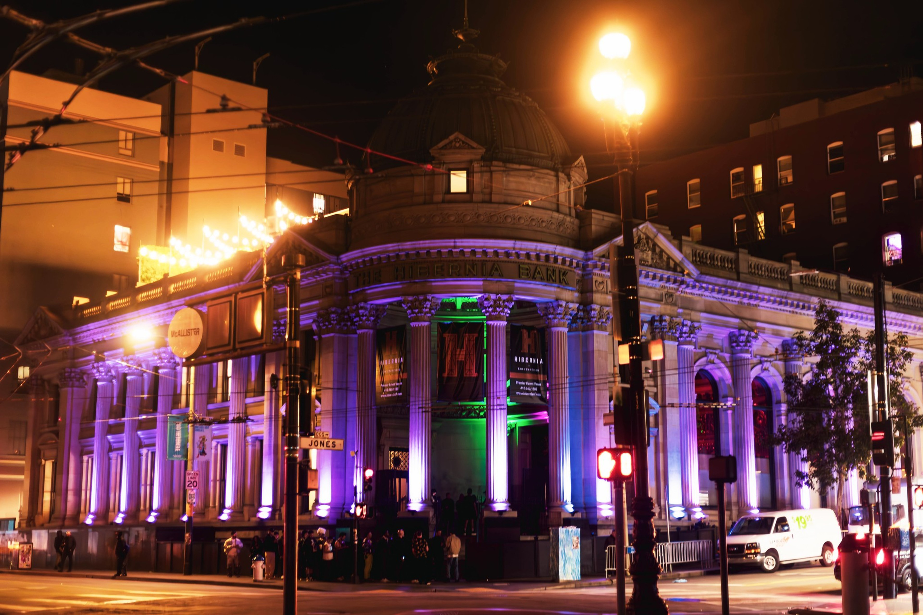 A well-lit, historic bank building at night with columns is adorned with purple and green lights. People are gathered at the entrance near a "McAllister" street sign.