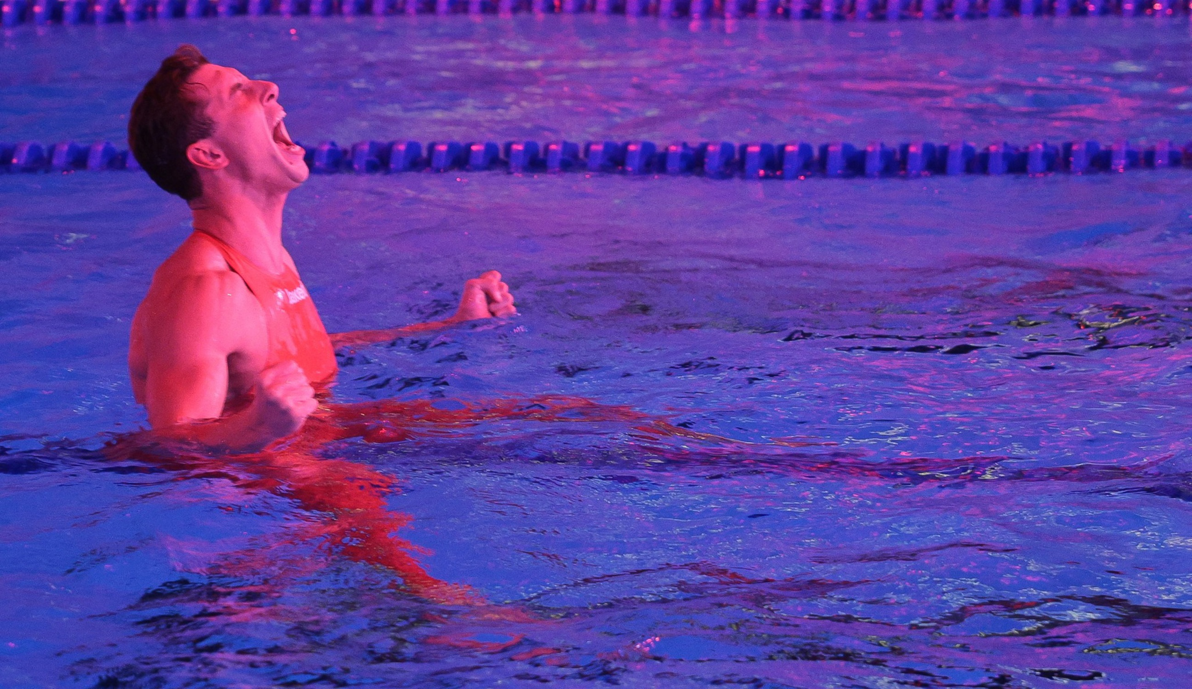 A swimmer in a red suit clenches his fists and shouts triumphantly while floating in a pool lane under purple lighting.