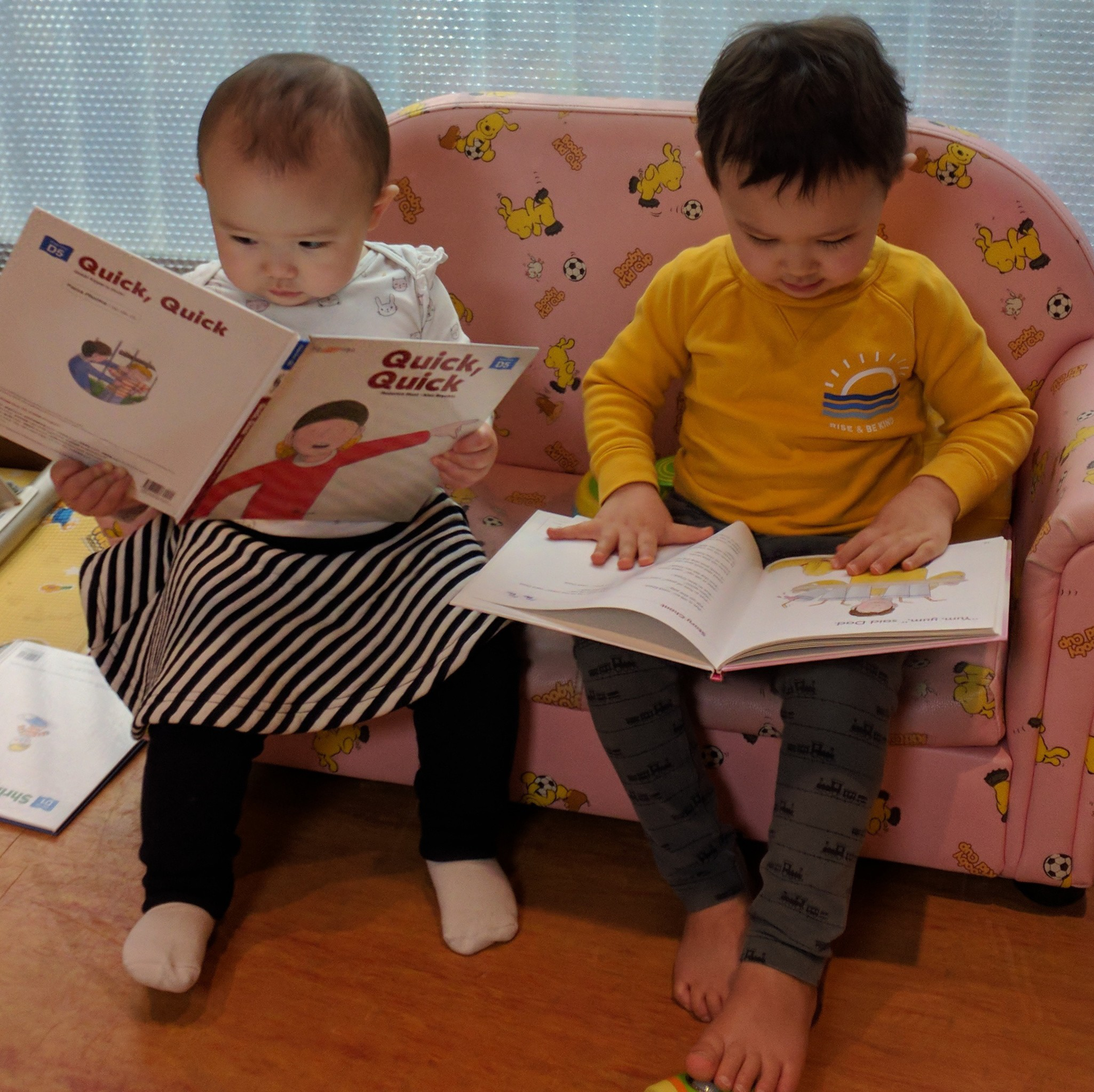 Two toddlers sit on a pink couch with cartoon dog prints, each focused on reading a book. One wears a striped skirt and the other a yellow shirt.