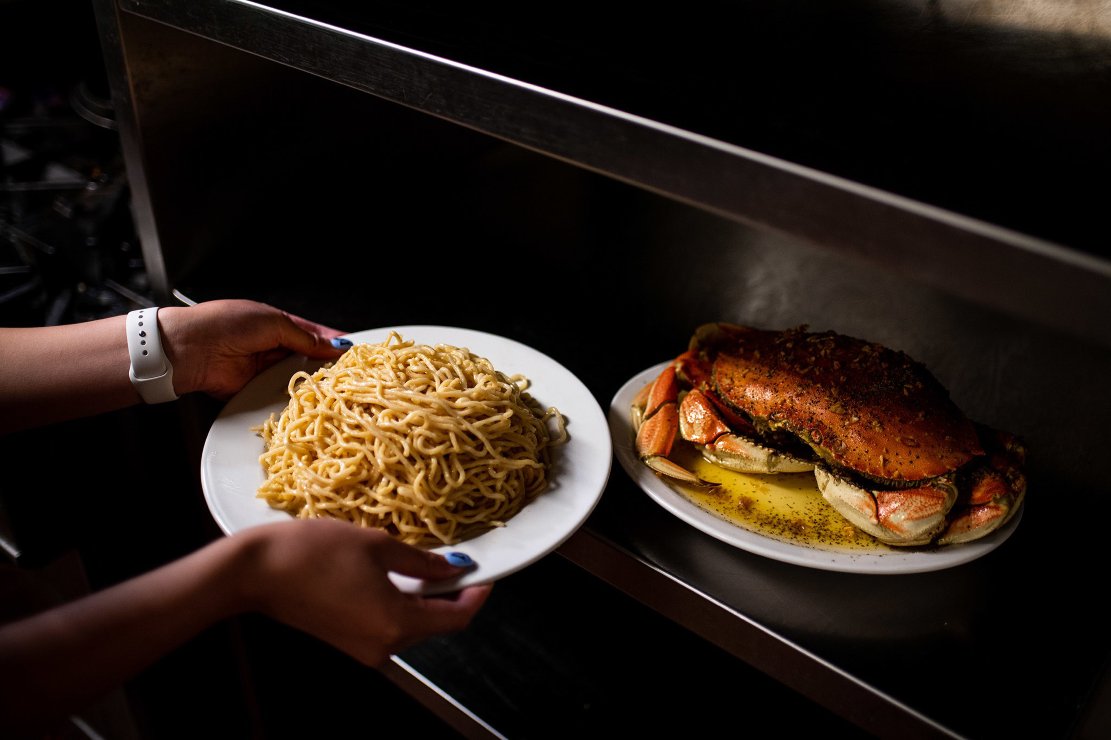 A person holds a plate of cooked noodles near a plate with a large cooked crab in seasoned butter on a metal shelf.