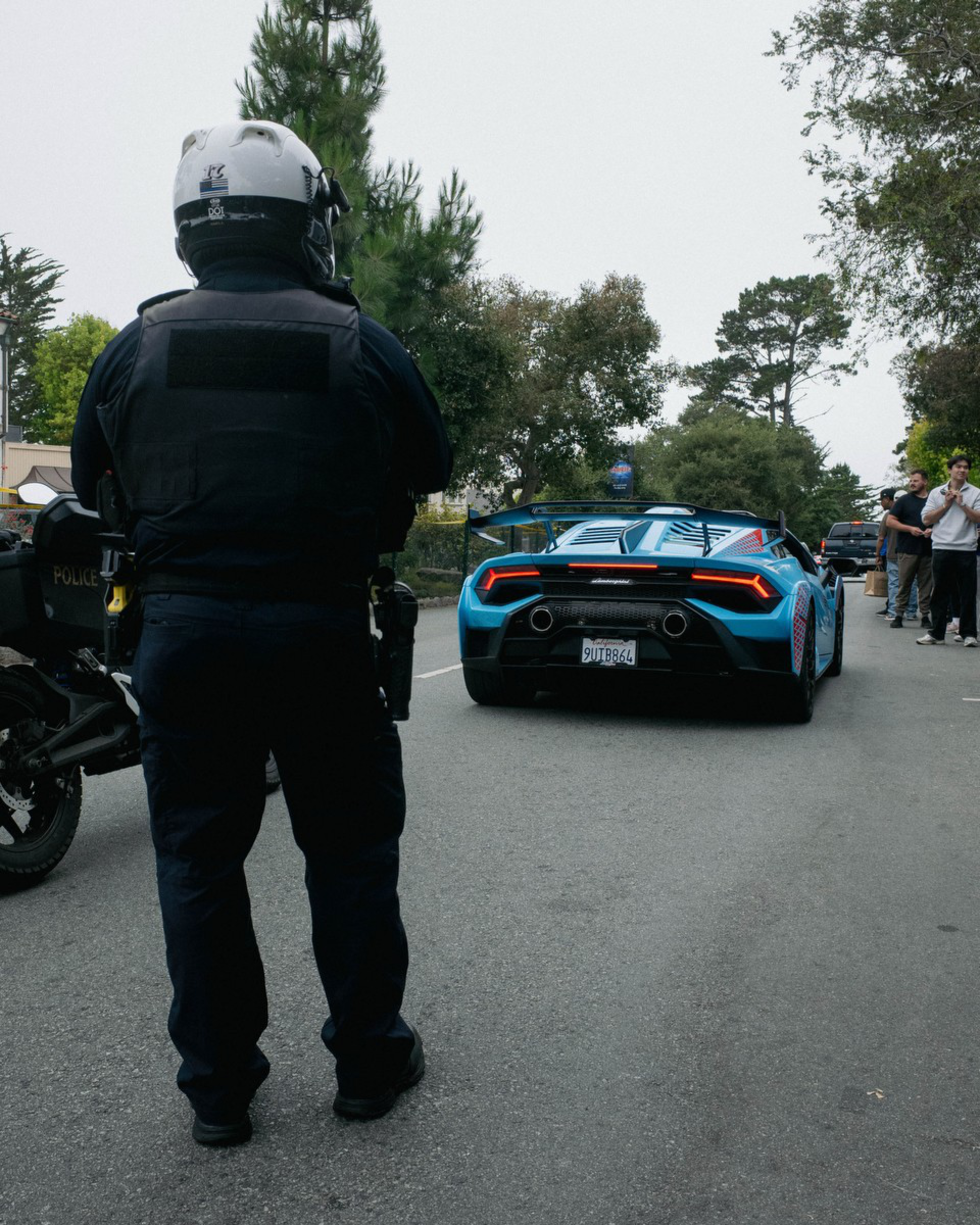 A police officer in gear stands by a motorcycle as a blue Lamborghini sports car drives on a road with people watching from the side.