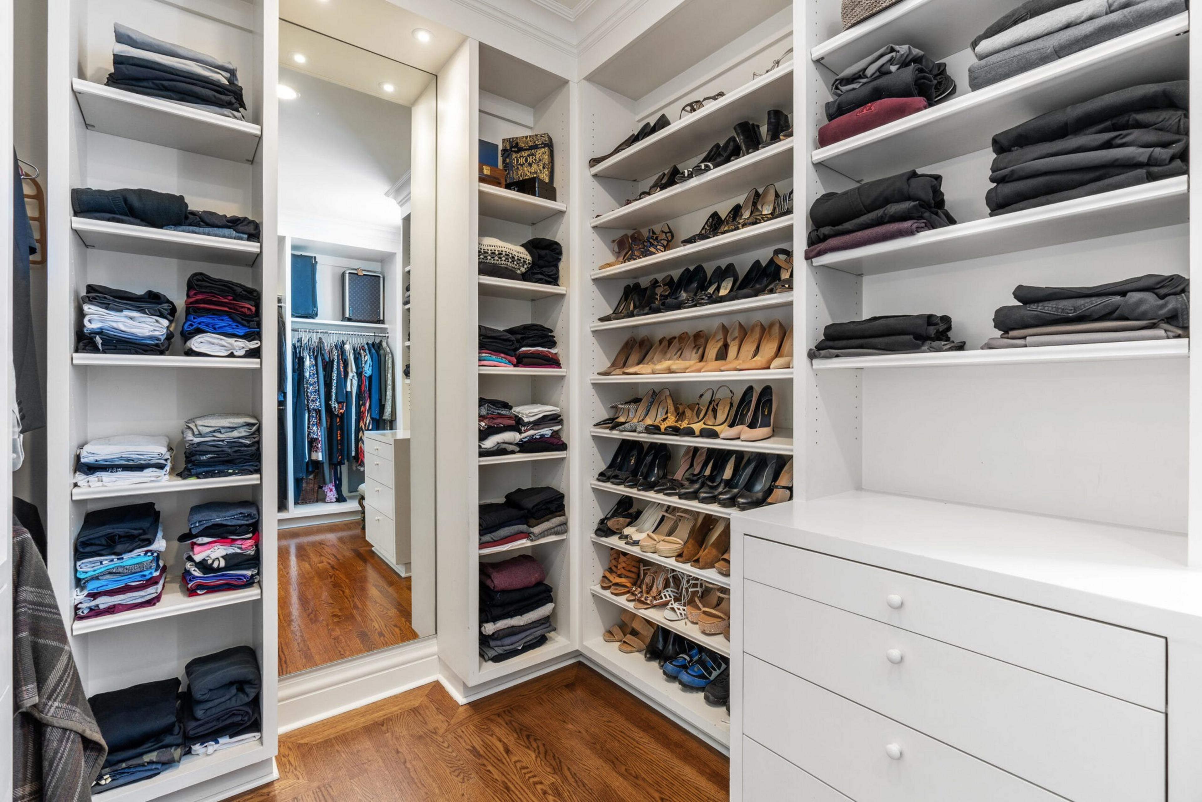 White built-in shelves and drawers hold neatly folded clothes and rows of shoes on wooden floors, with a tall mirror reflecting clothes hanging in another closet.