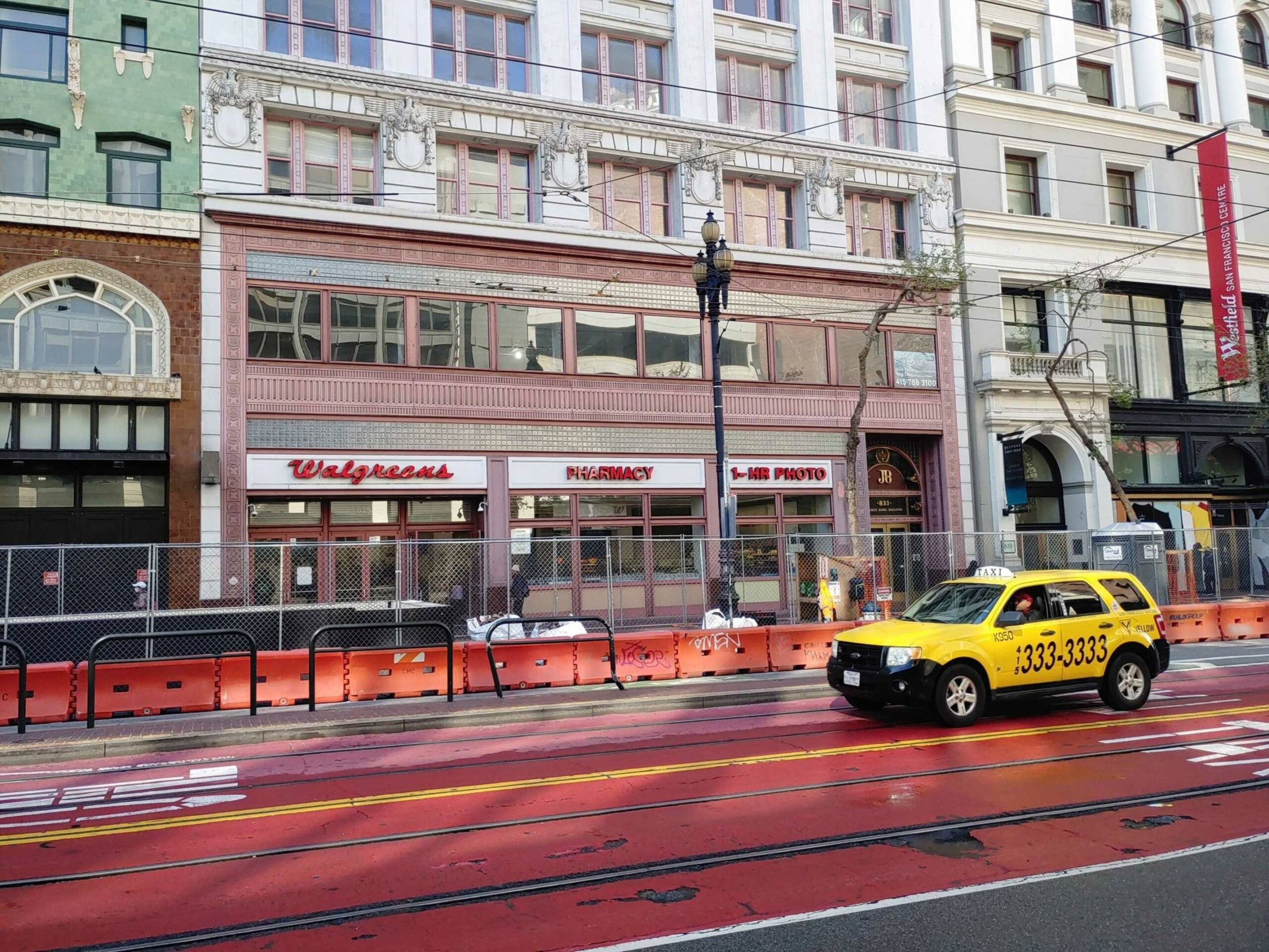A yellow taxi drives past a Walgreens store with red and white signage, behind orange construction barriers lining the sidewalk.