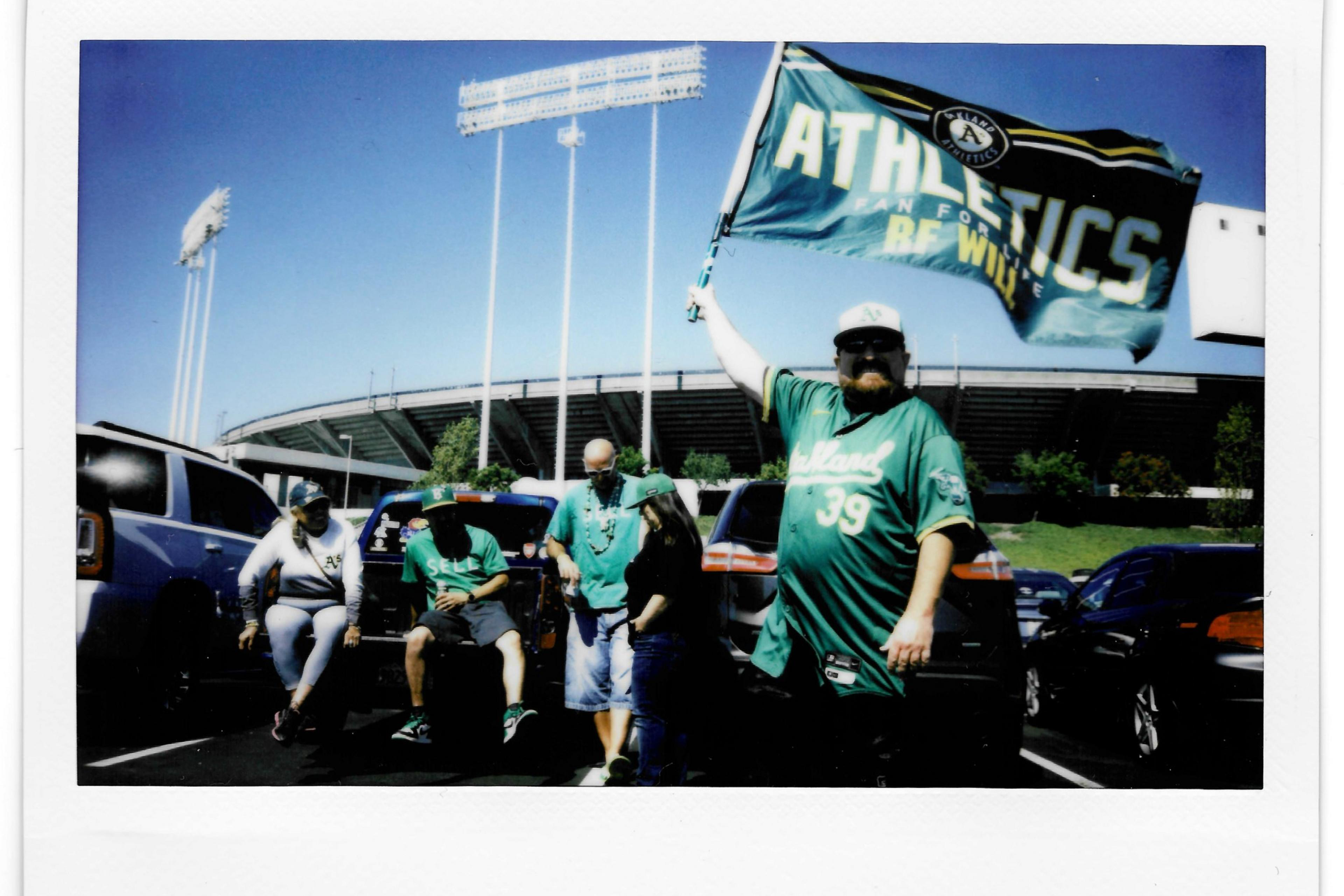 A group of fans in Oakland Athletics attire stands and sits by cars in a parking lot, with one man holding a large Athletics flag. The stadium is visible in the background.