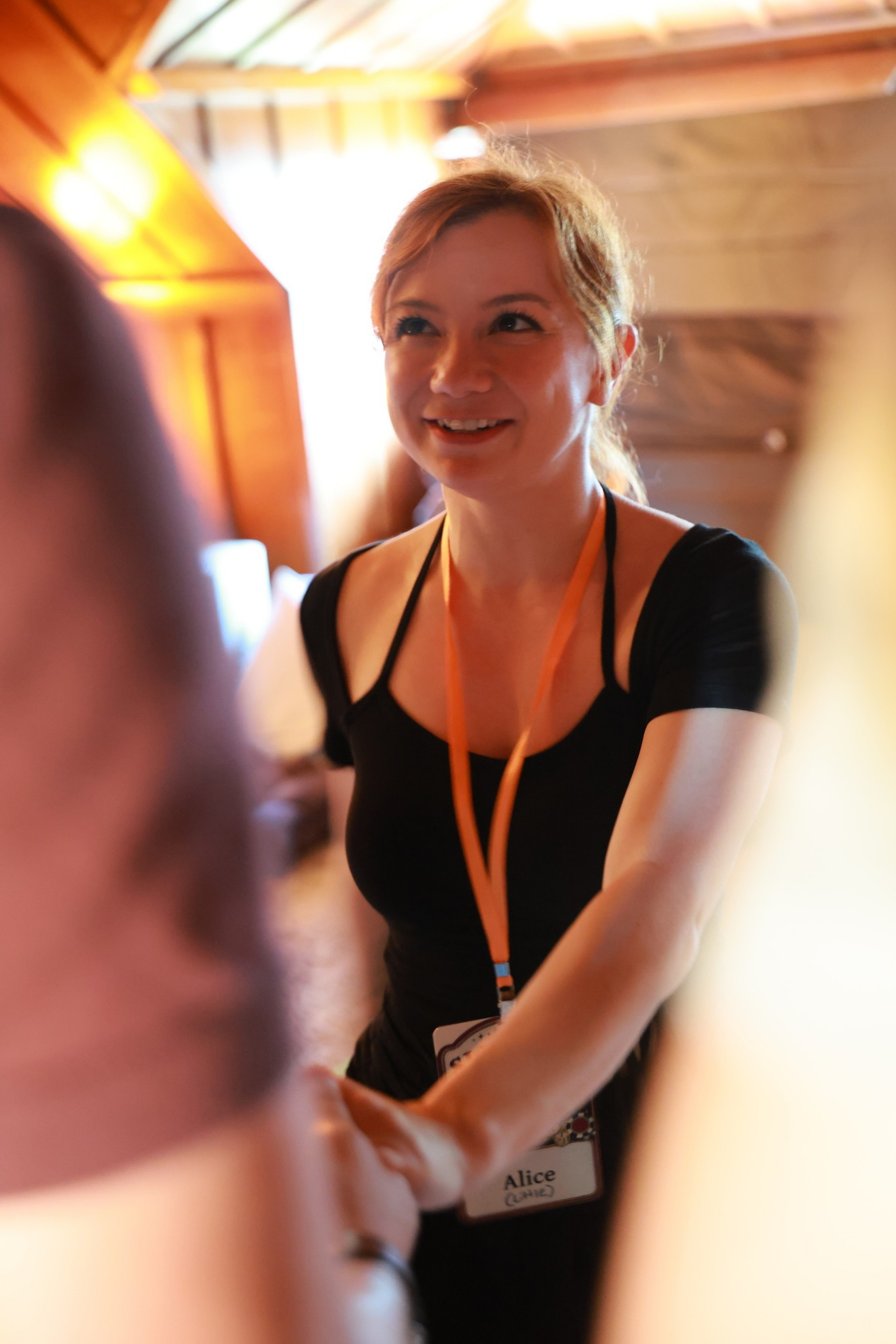 A woman with light hair, wearing a black top and an orange lanyard with a name tag that reads “Alice,” is smiling and shaking hands indoors.