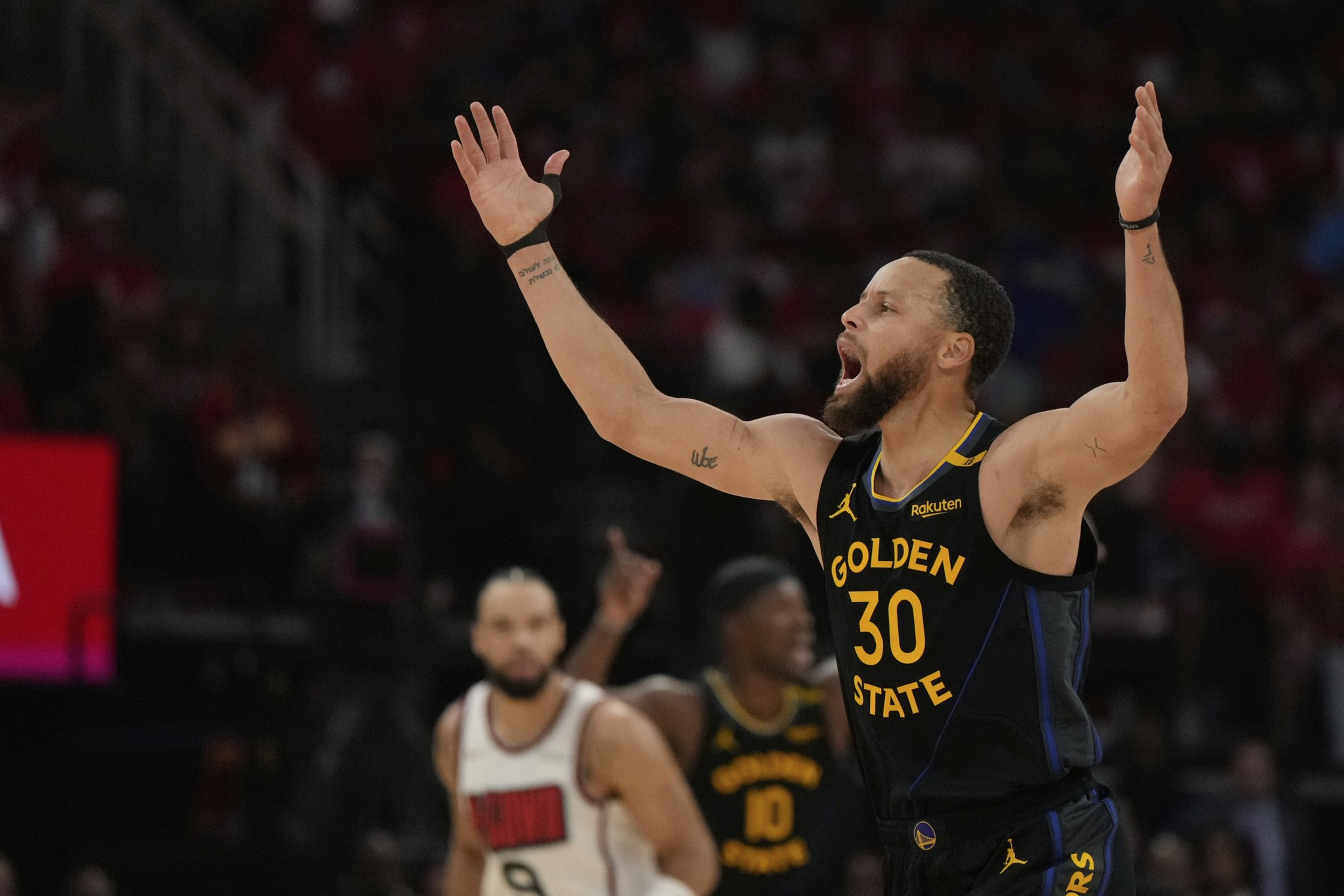 A basketball player in a Golden State Warriors uniform (#30) raises his arms, appearing to shout. Other players are blurred in the background on the court.