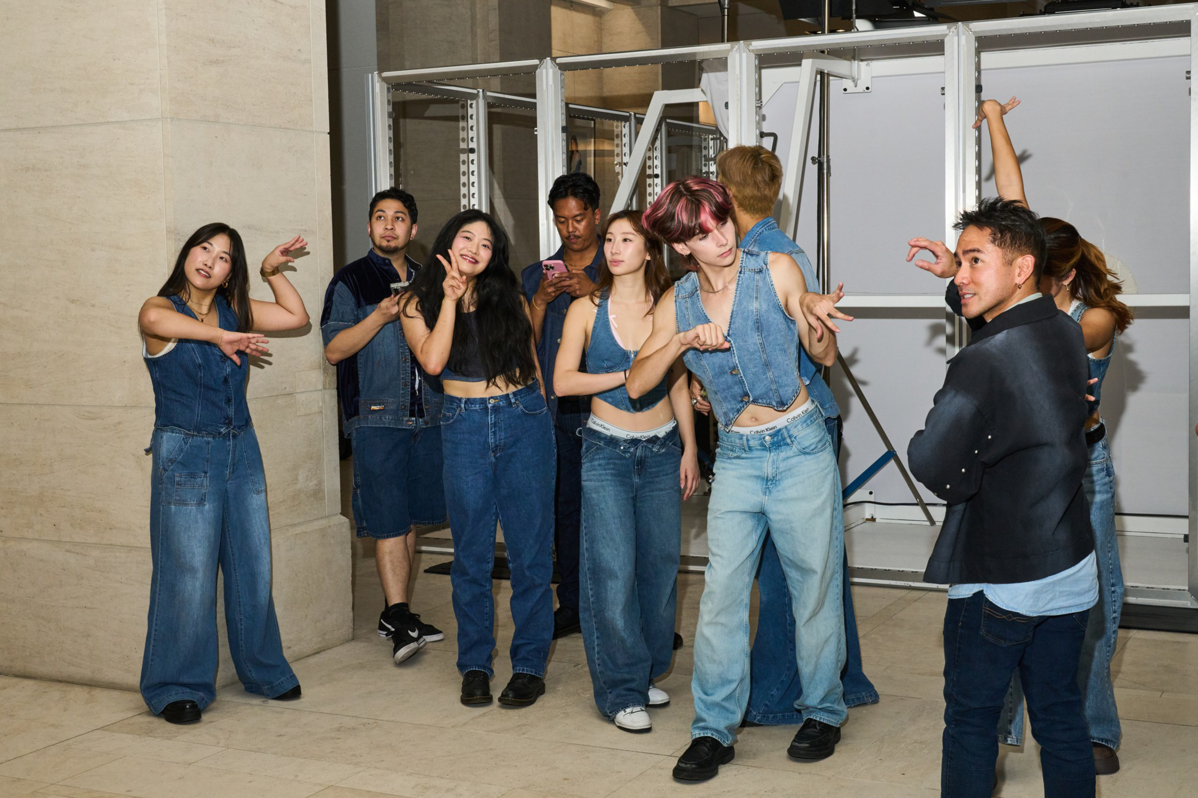 Eight people in denim outfits stand indoors, some posing with hand gestures, while one person in a black jacket faces them with a smile.
