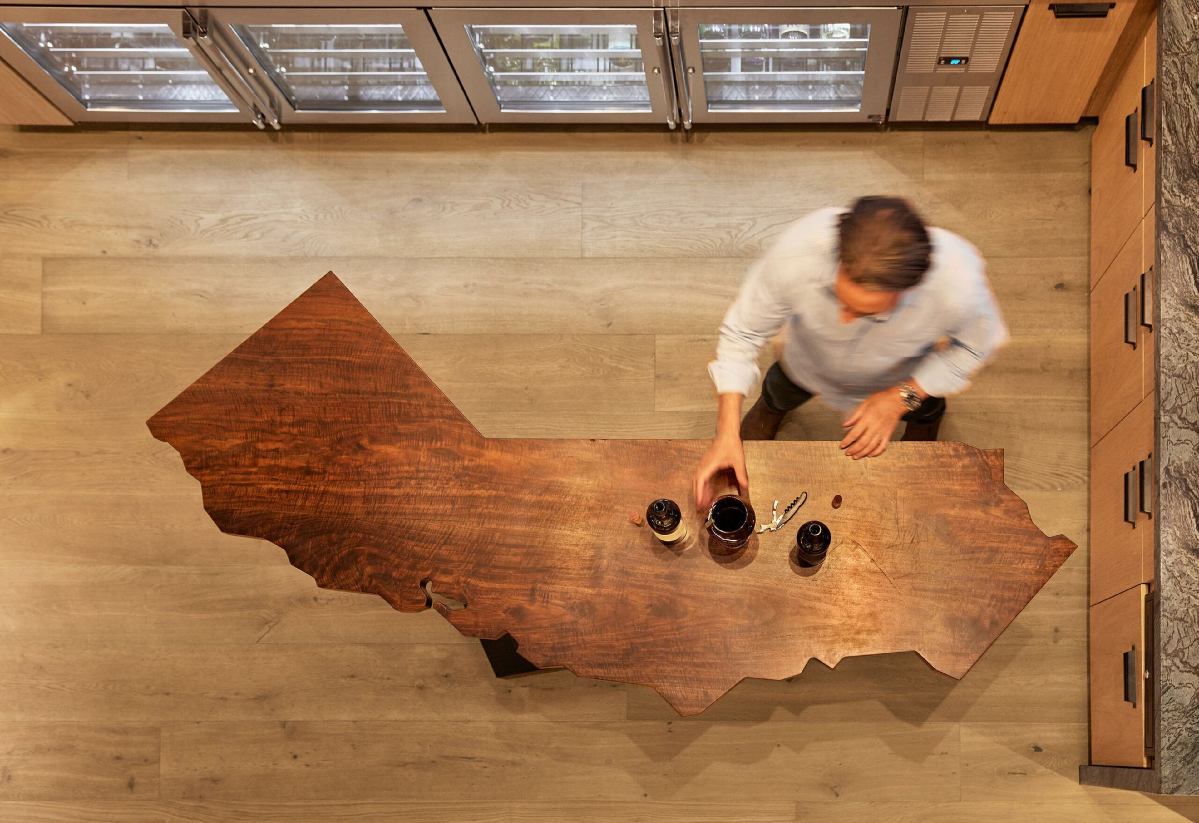 A person stands at a wooden table shaped like a U.S. state, holding a glass of red wine with two bottles nearby, in a room with wooden floors and cabinets.