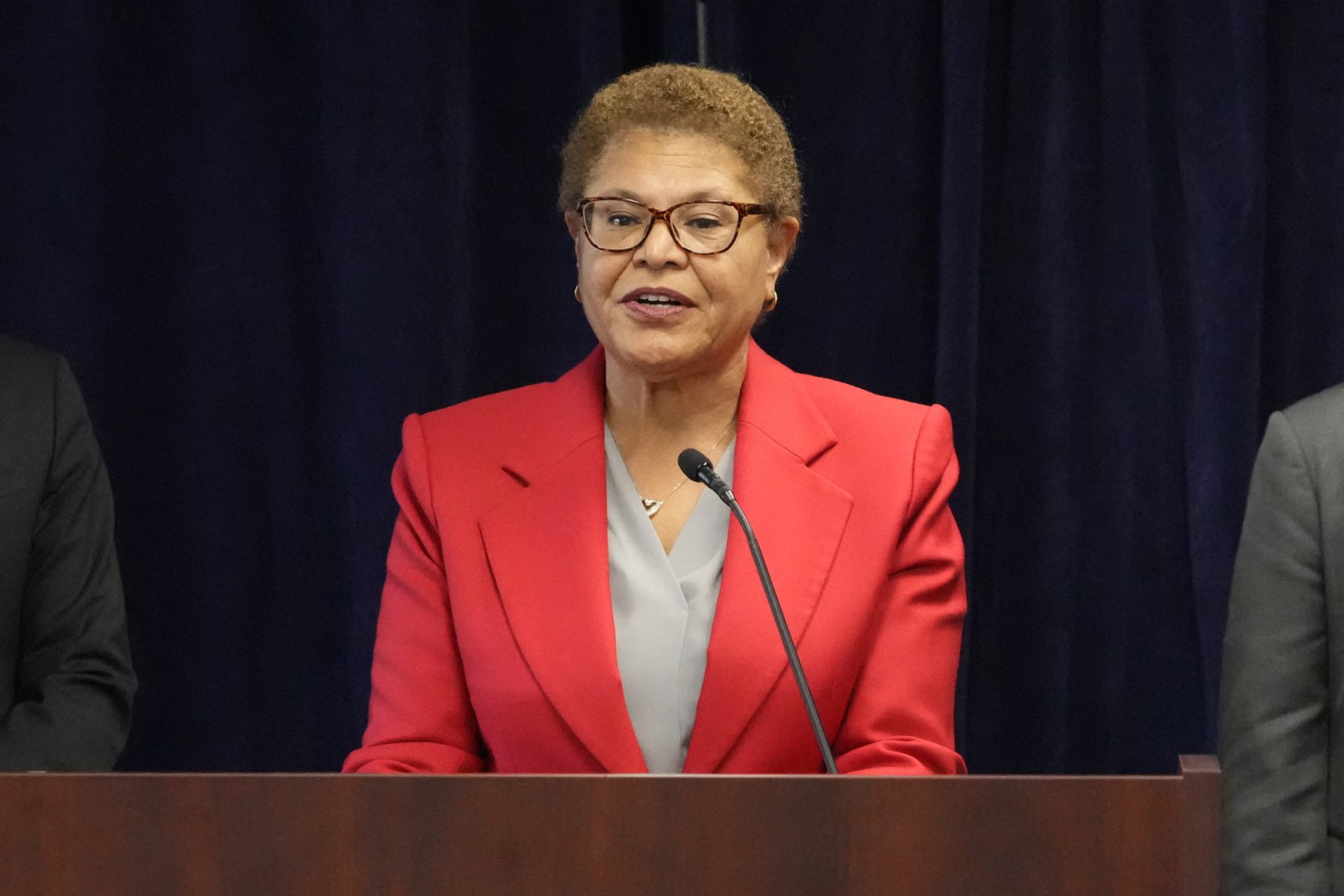 A woman wearing glasses and a red blazer stands behind a wooden podium, speaking into a microphone against a dark background.