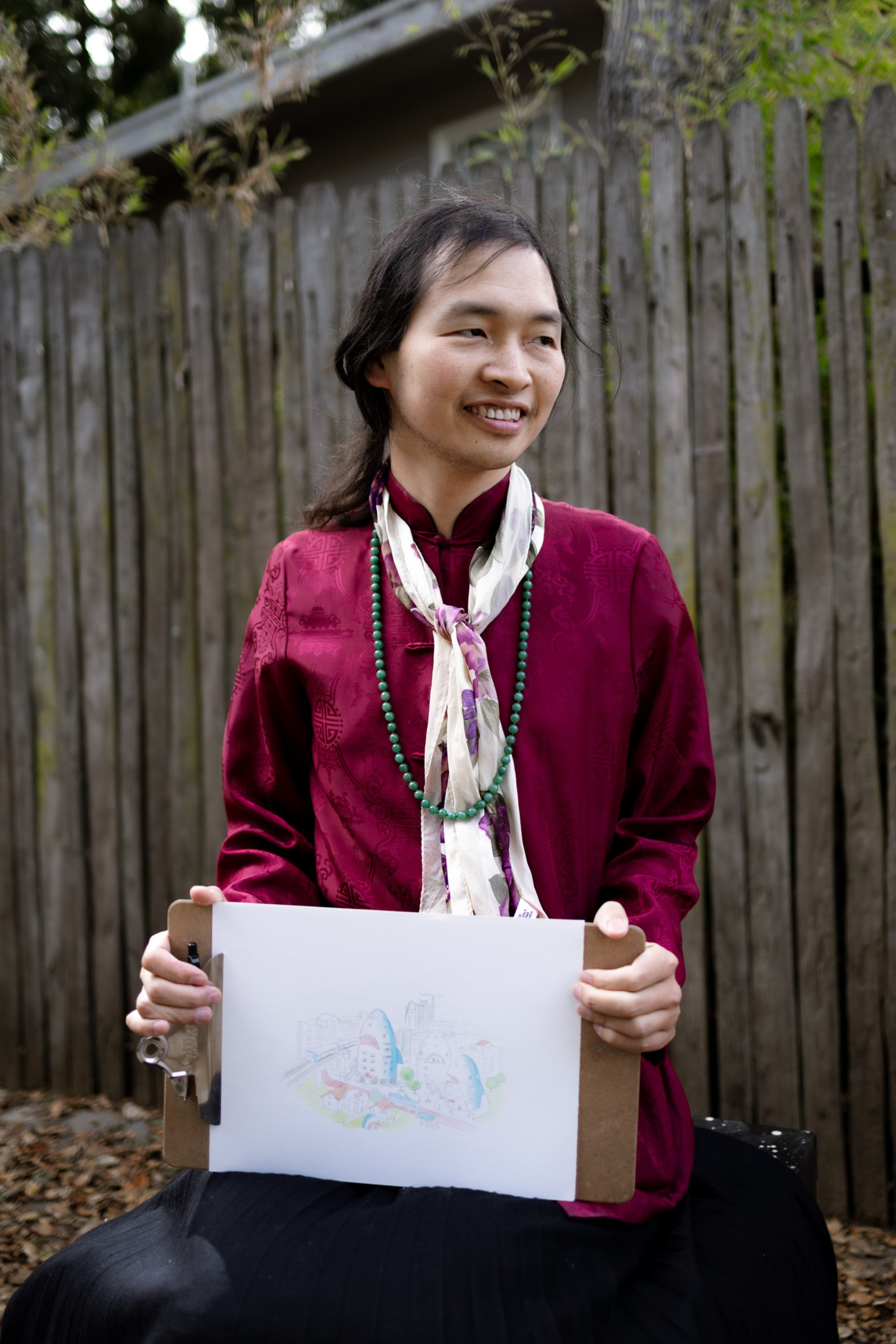 A person wearing a burgundy traditional garment and a floral scarf holds a clipboard with a colorful drawing, sitting outdoors in front of a wooden fence.