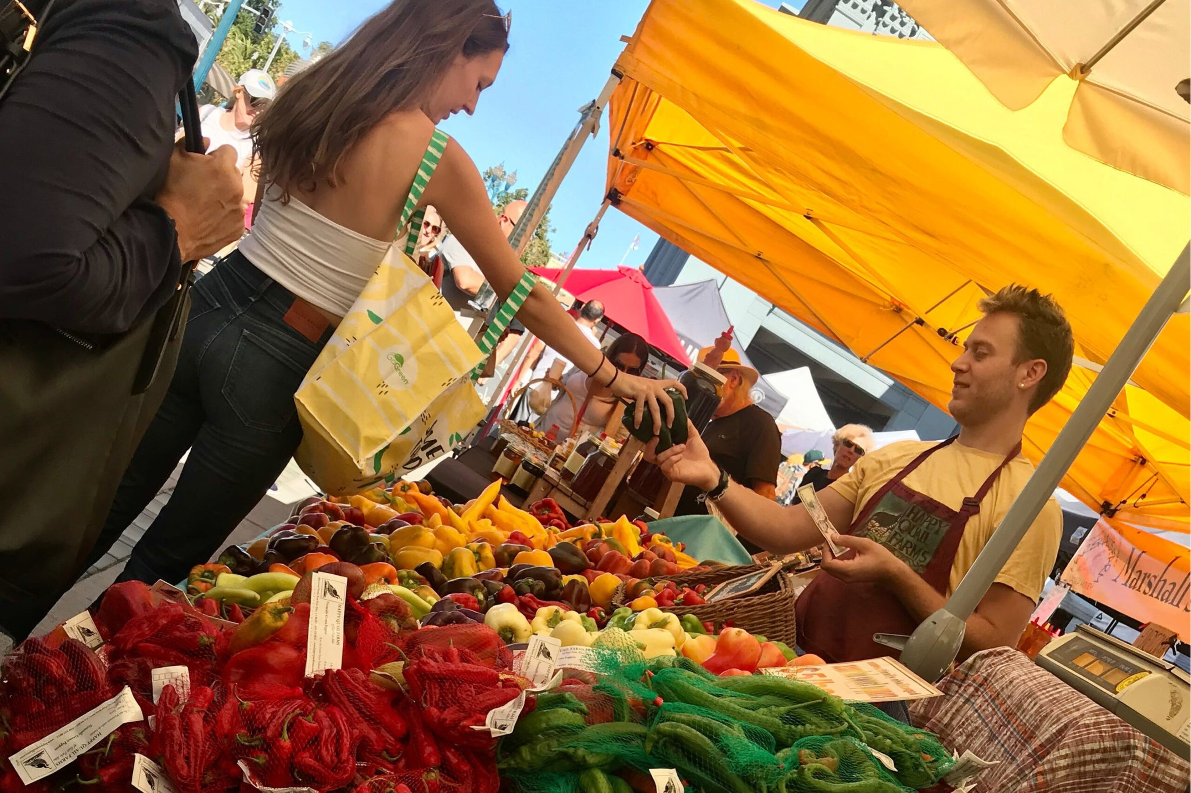 A woman is buying fresh produce from a vendor at an outdoor market with colorful fruits and vegetables under yellow tents.