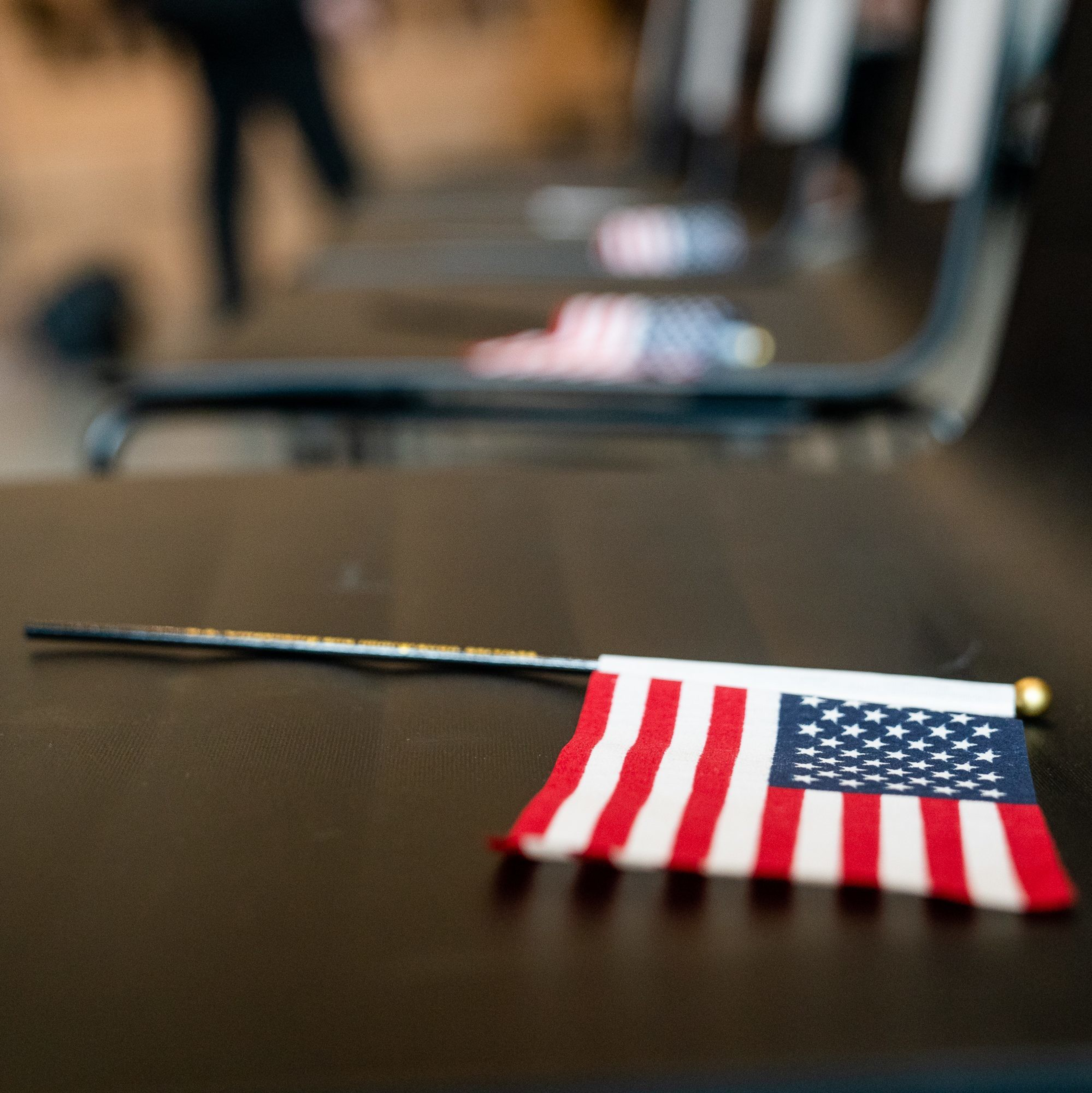 A close-up image of a small American flag on a stick resting on a row of empty black chairs, suggesting a formal setting.