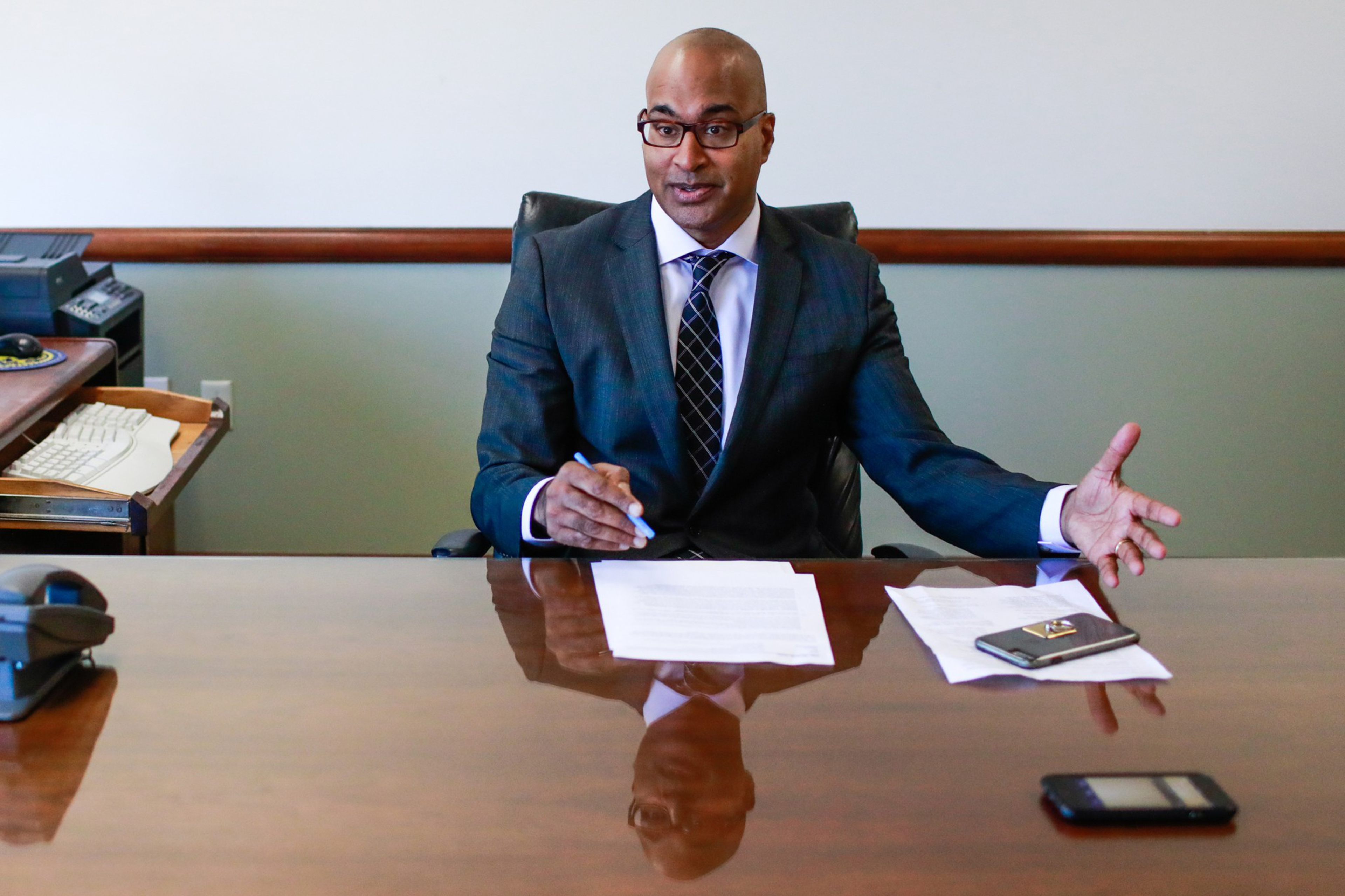 A person in a suit sits at a wooden desk with papers, a pen, and two phones. They're gesturing with one hand, looking engaged in conversation.