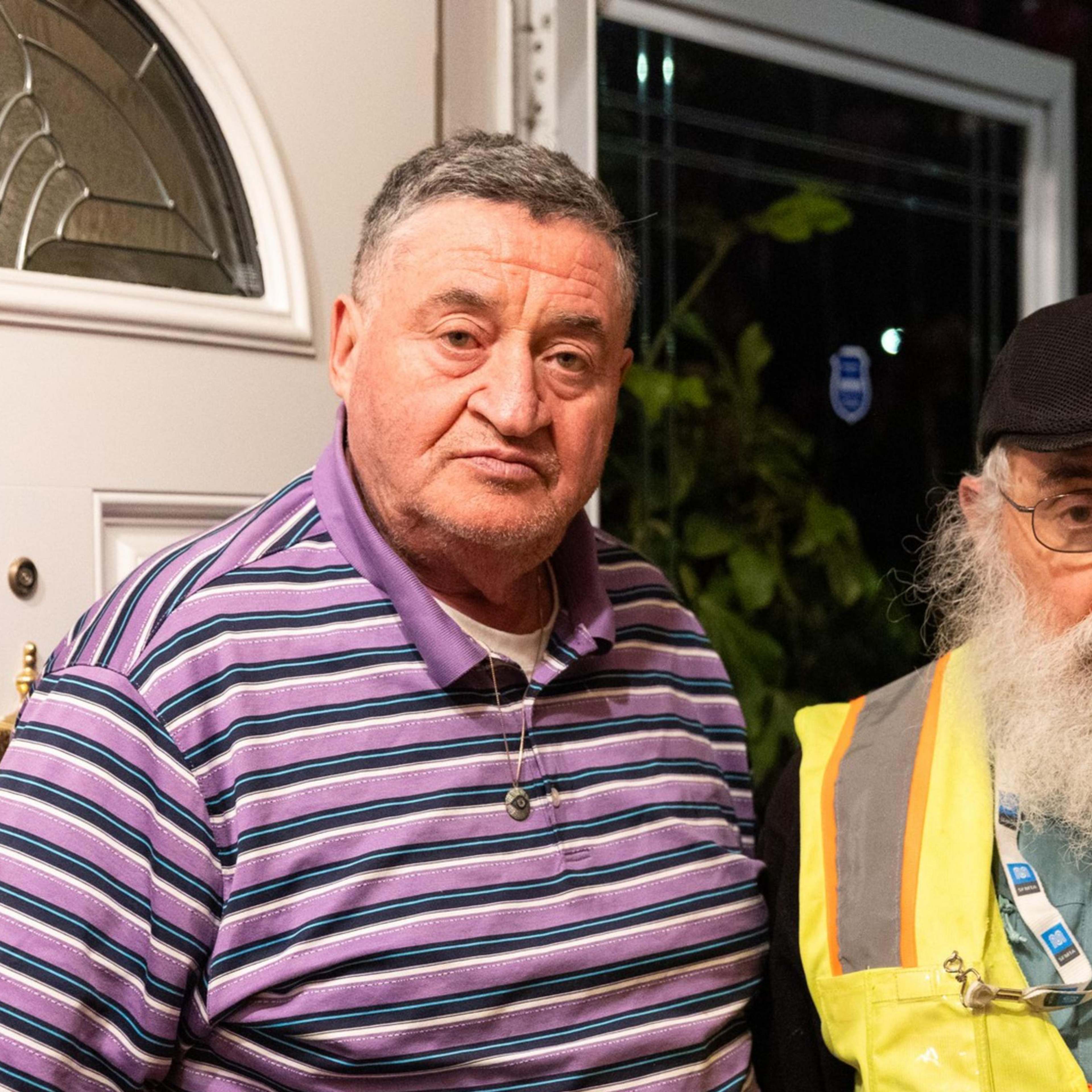 Two older men stand at a doorway. One wears a purple striped polo, and the other dons a black cap with a yellow reflective vest.