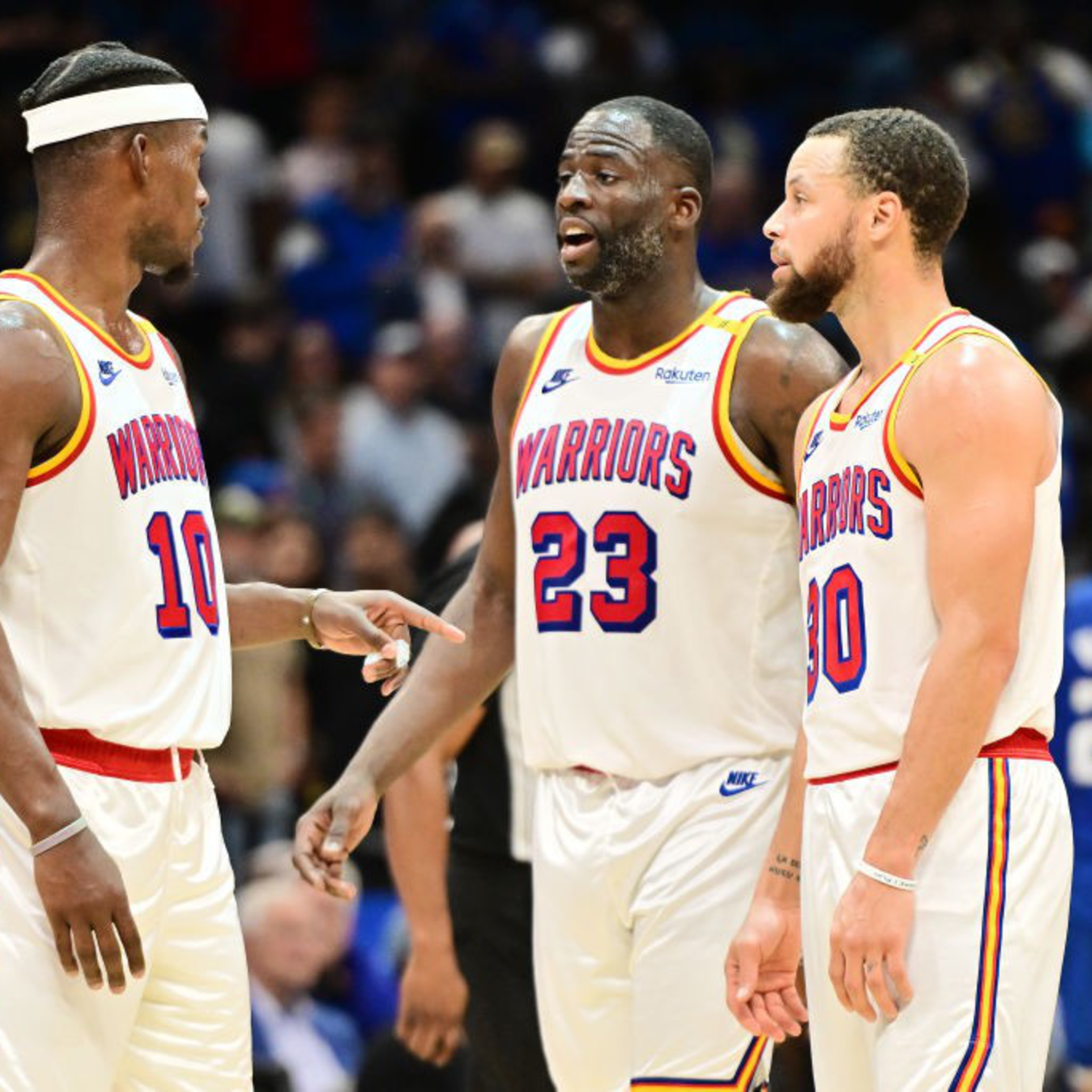 Three basketball players in white "Warriors" jerseys are talking on the court. One is wearing a headband, and number 23 is speaking in the middle.