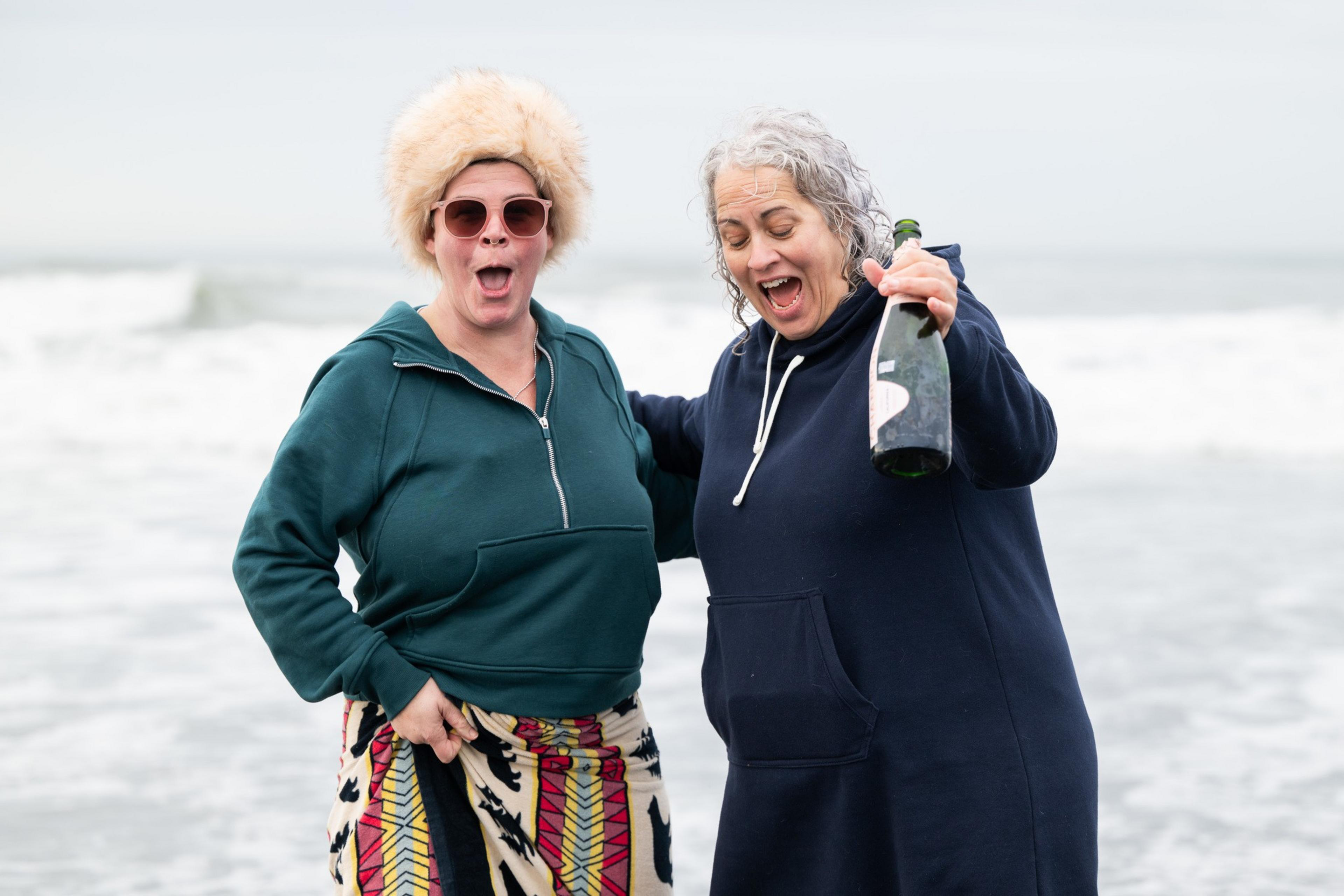 Two people stand by the beach, smiling exuberantly. One wears a furry hat and sunglasses, the other holds a bottle. Both wear hooded outfits.