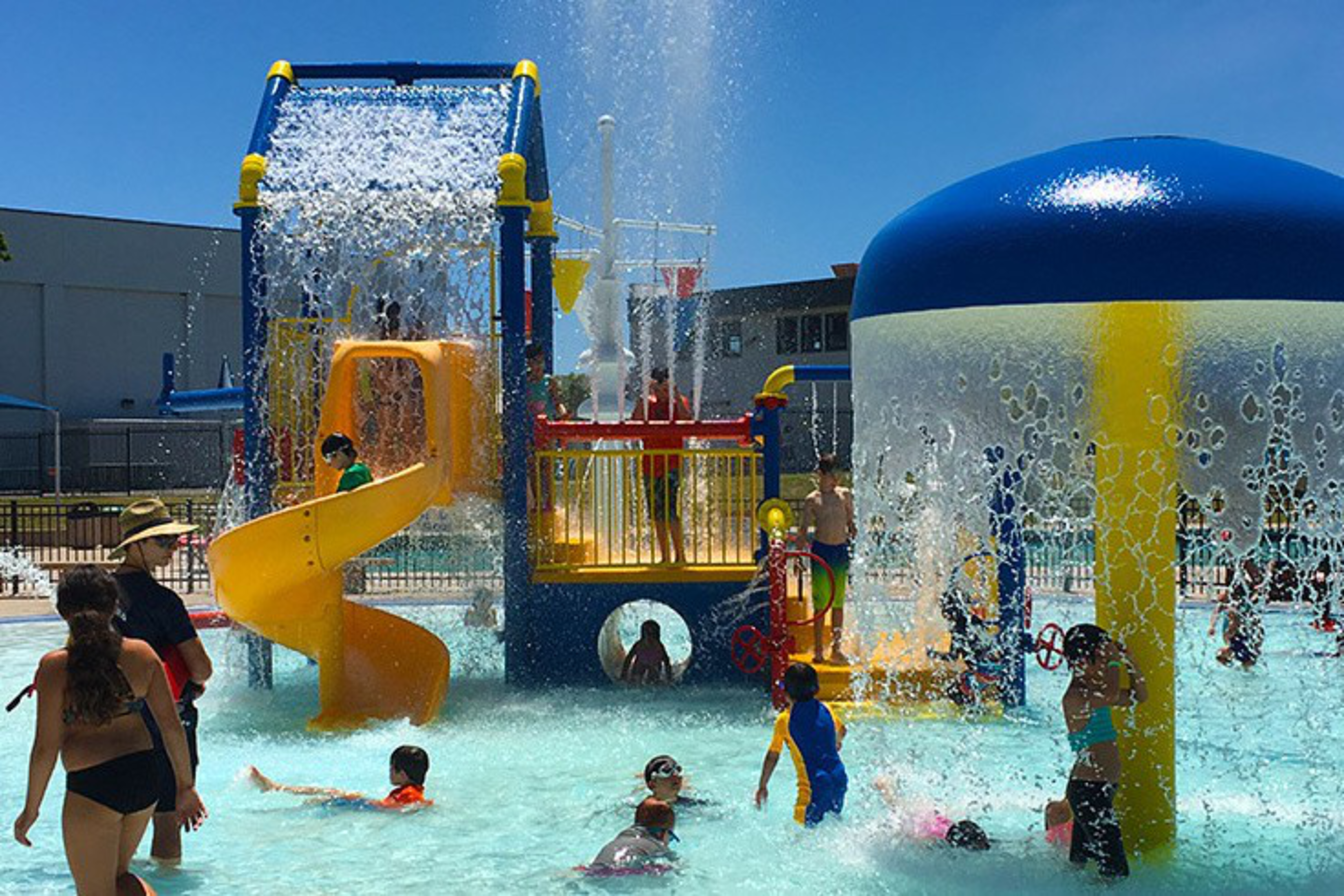Children and adults play in a water park with slides, splash structures, and cascading water under a clear blue sky.