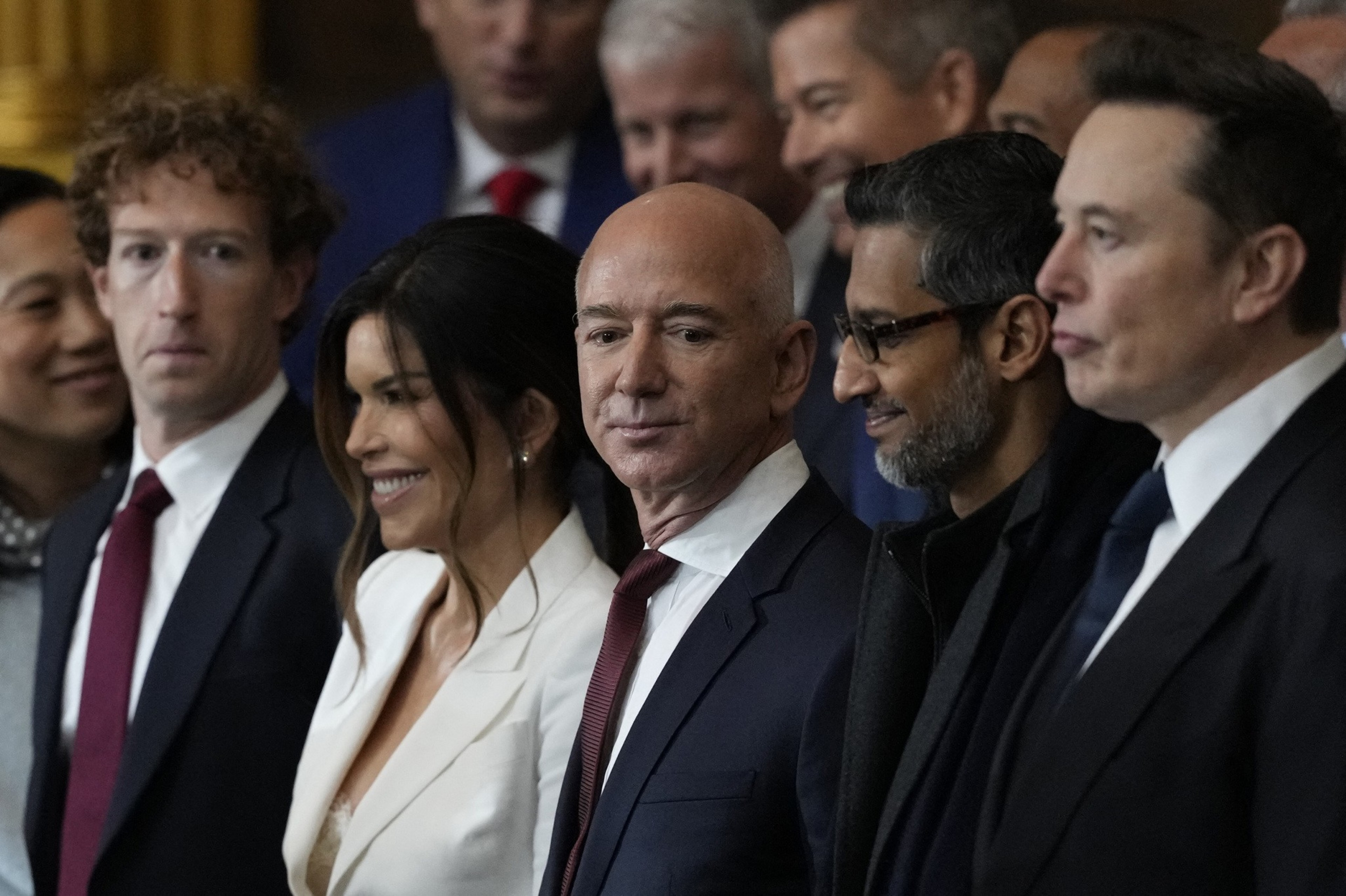 A group of people dressed in formal attire are standing closely together. They appear to be at an event or gathering, with some smiling and others looking engaged.
