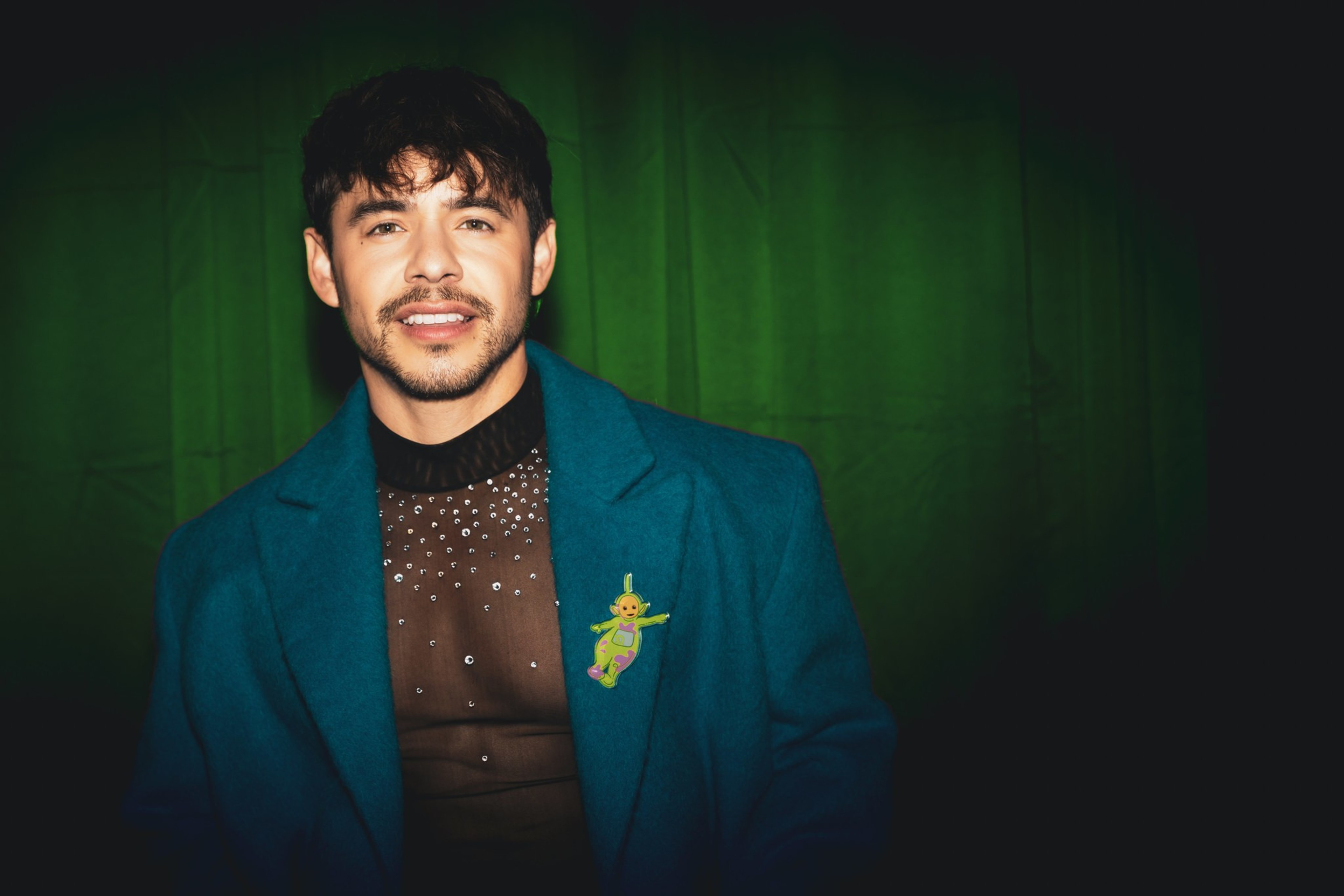Singer David Archuleta stands against a dark green background, wearing a blue coat over a black, pearl-studded shirt. A colorful pin in the shape of an alien is on the coat.