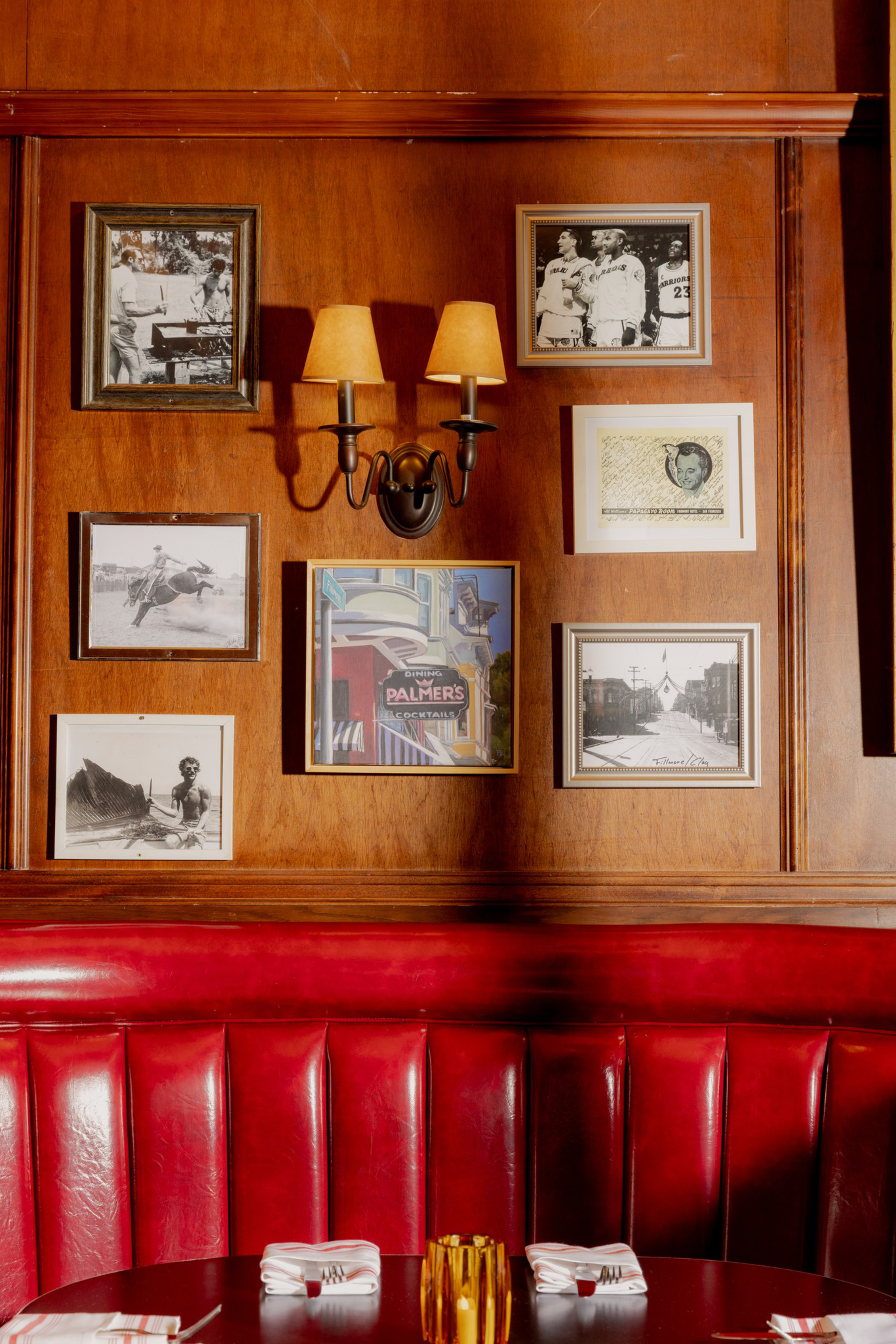 A wood-paneled wall above a red leather booth features six framed photos, a central colorful sign, and a double wall sconce with two lit lamps. Two tables are set below.