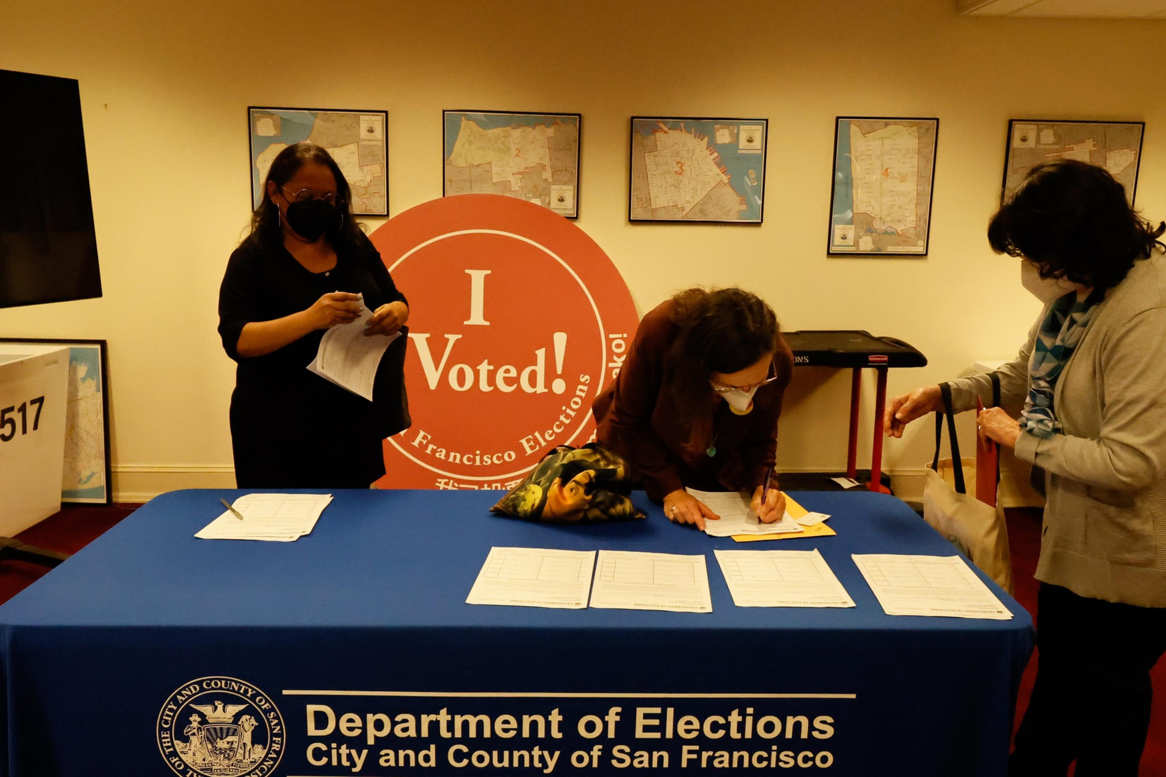 Three people at a Department of Elections table in San Francisco, one signing papers, with an "I Voted!" sign and maps on the wall behind them.