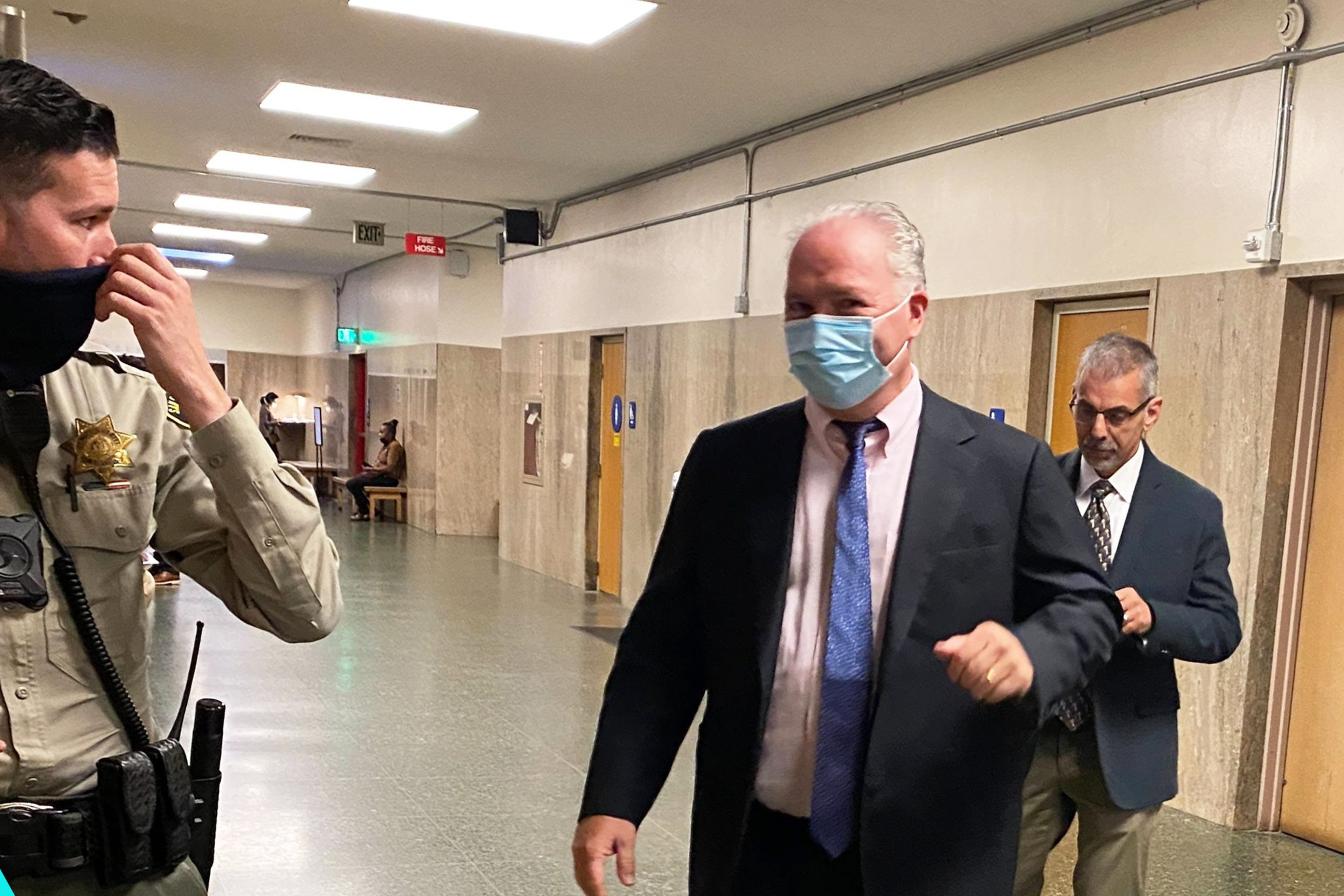 A man in a suit and blue mask walks through a hallway with another man in a suit behind him, while a uniformed officer touches his mask on the left.