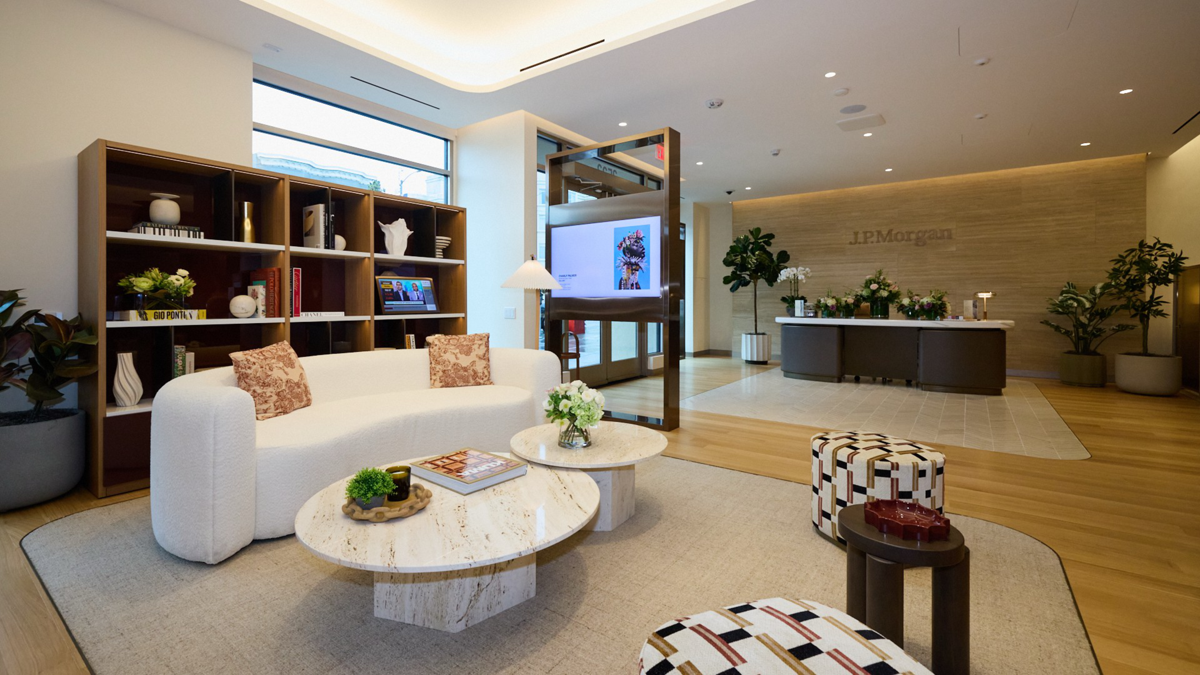 A modern office lounge with a white curved sofa, two round marble tables, patterned stools, potted plants, a bookshelf, and a reception desk labeled “J.P. Morgan.”