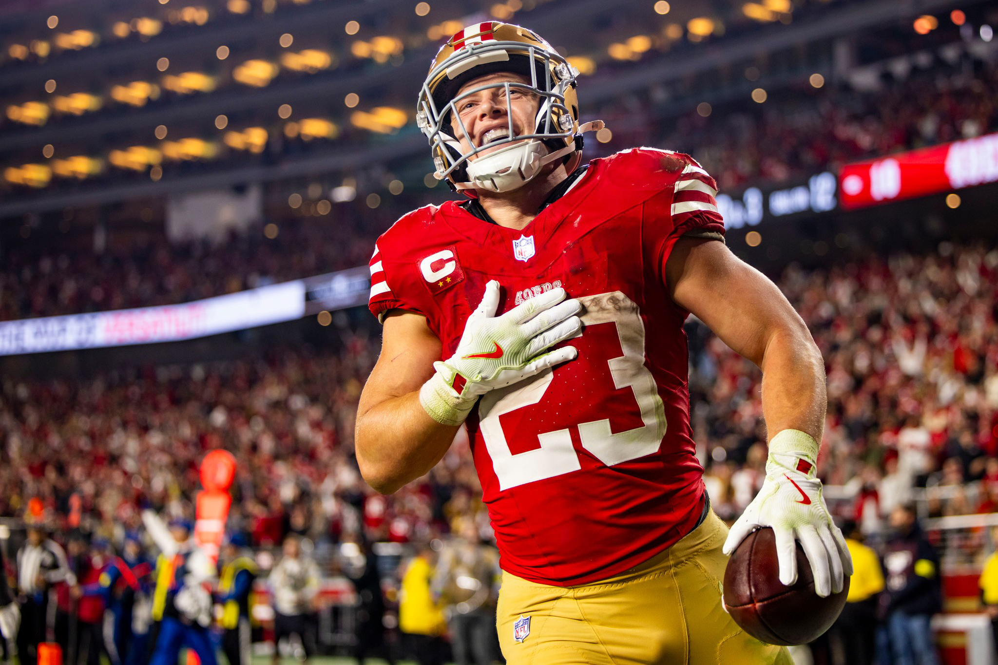 A football player wearing a red jersey with number 23, gold pants, and a helmet holds the ball and smiles with a crowd in the background.