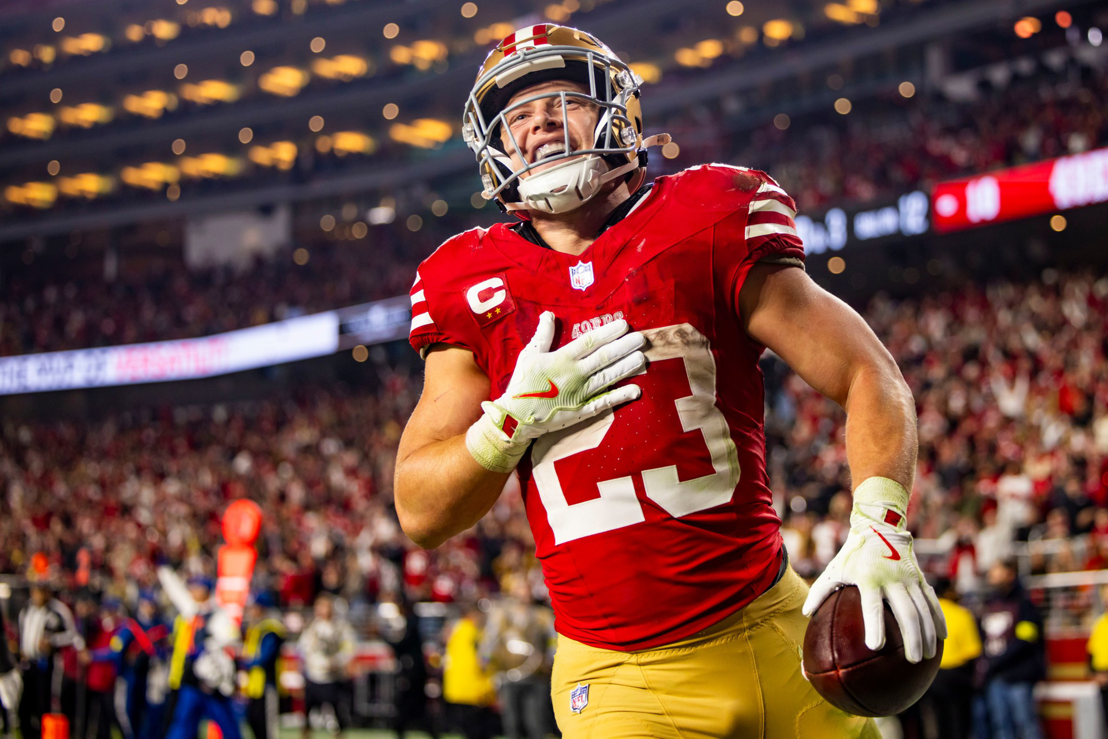 A football player wearing a red jersey with number 23, gold pants, and a helmet holds the ball and smiles with a crowd in the background.
