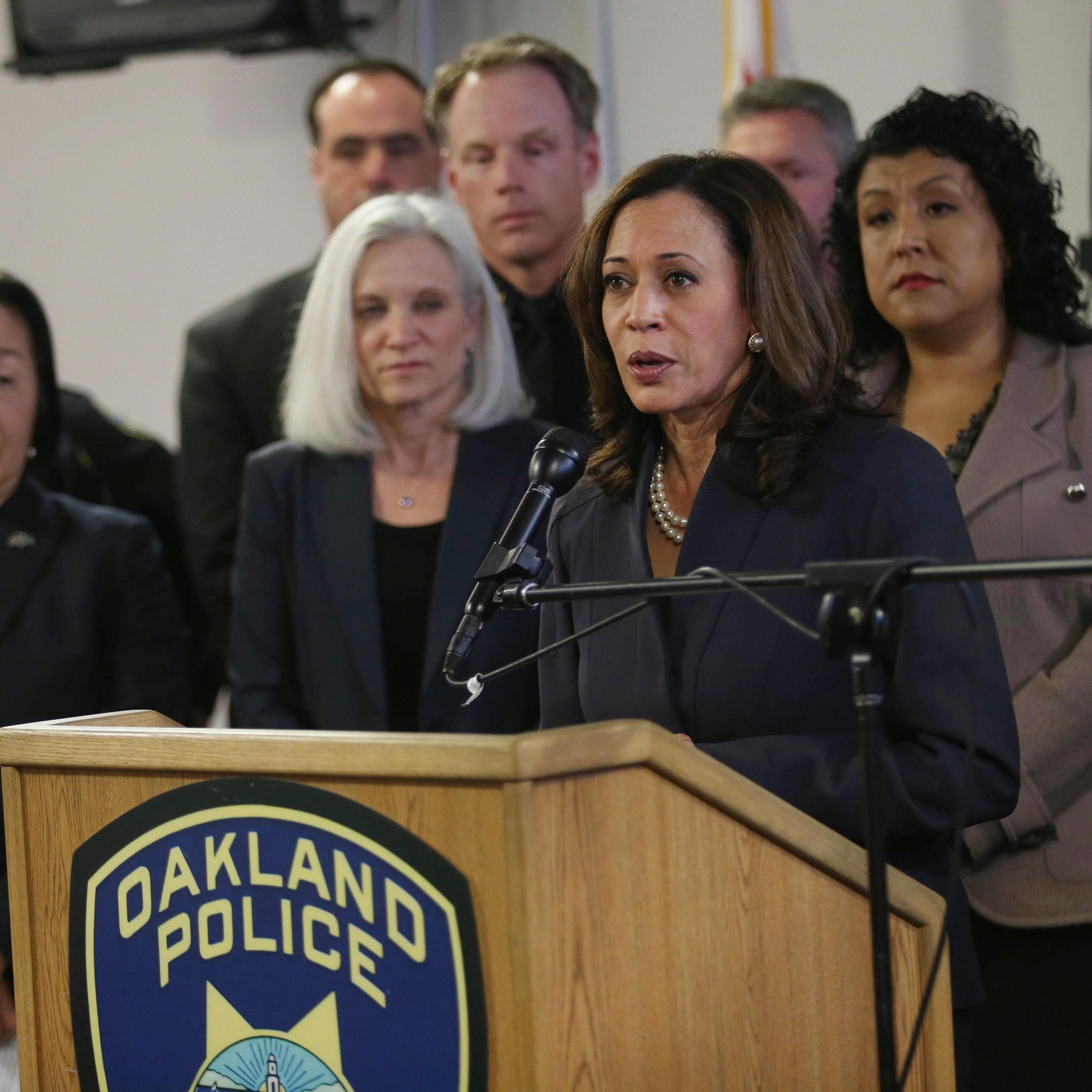 A woman speaks at a podium labeled &quot;Oakland Police,&quot; surrounded by five individuals who appear attentive in a formal setting. A microphone is positioned in front.
