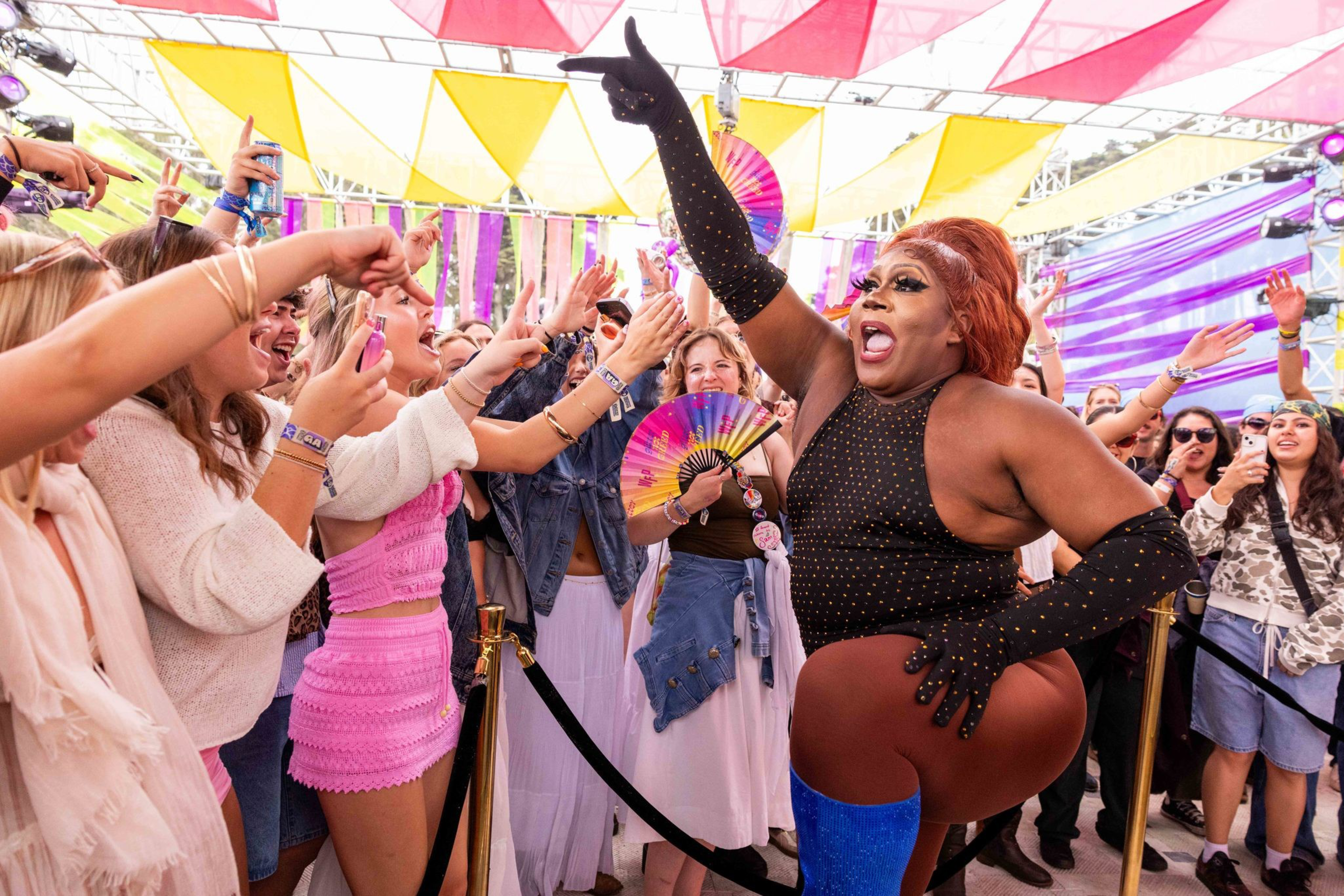 A performer in a black studded outfit and blue boots energizes a cheering crowd under colorful triangle flags at an outdoor event.