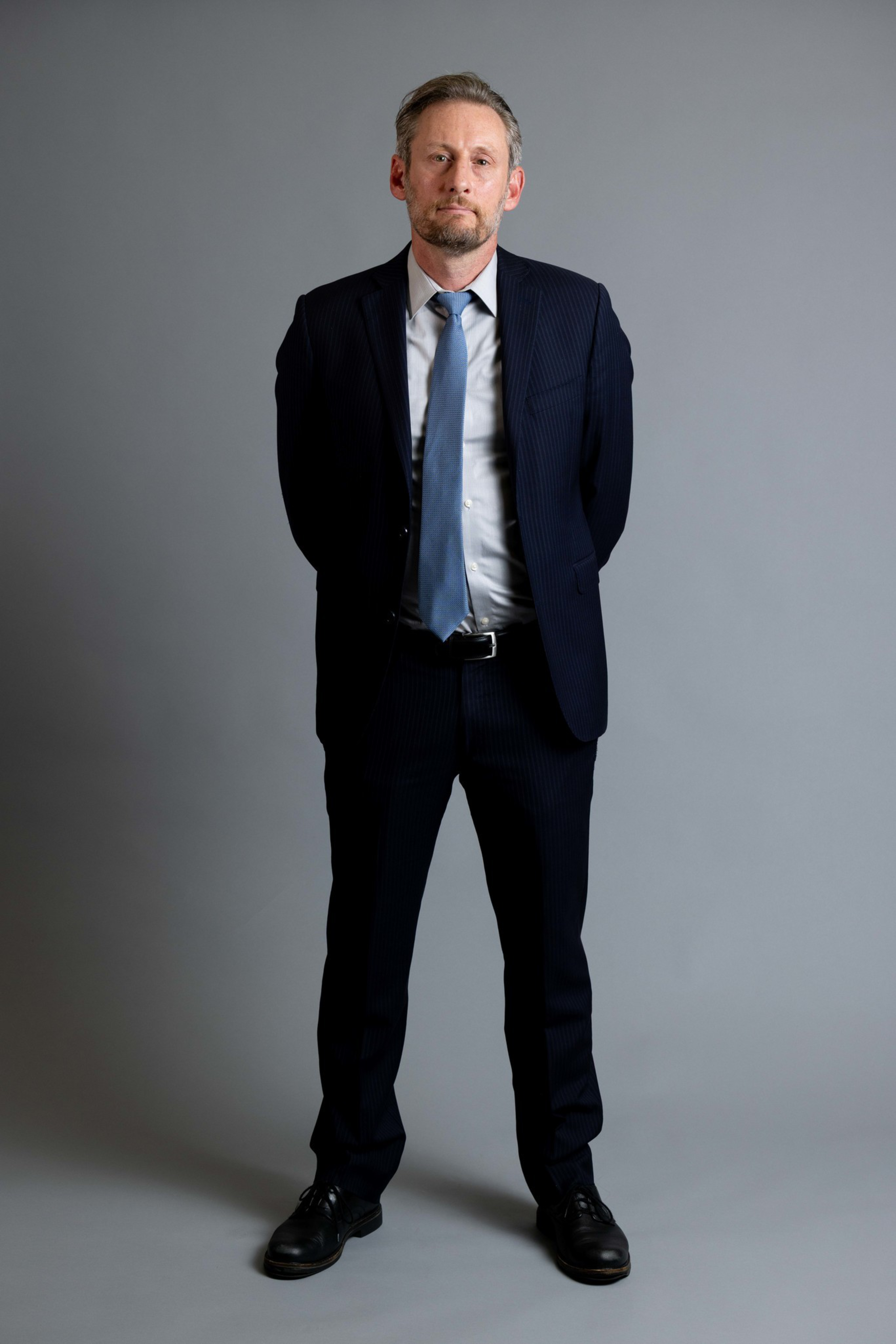 A man in a dark suit, light blue shirt, and blue tie stands with hands behind his back against a plain gray background, looking at the camera.