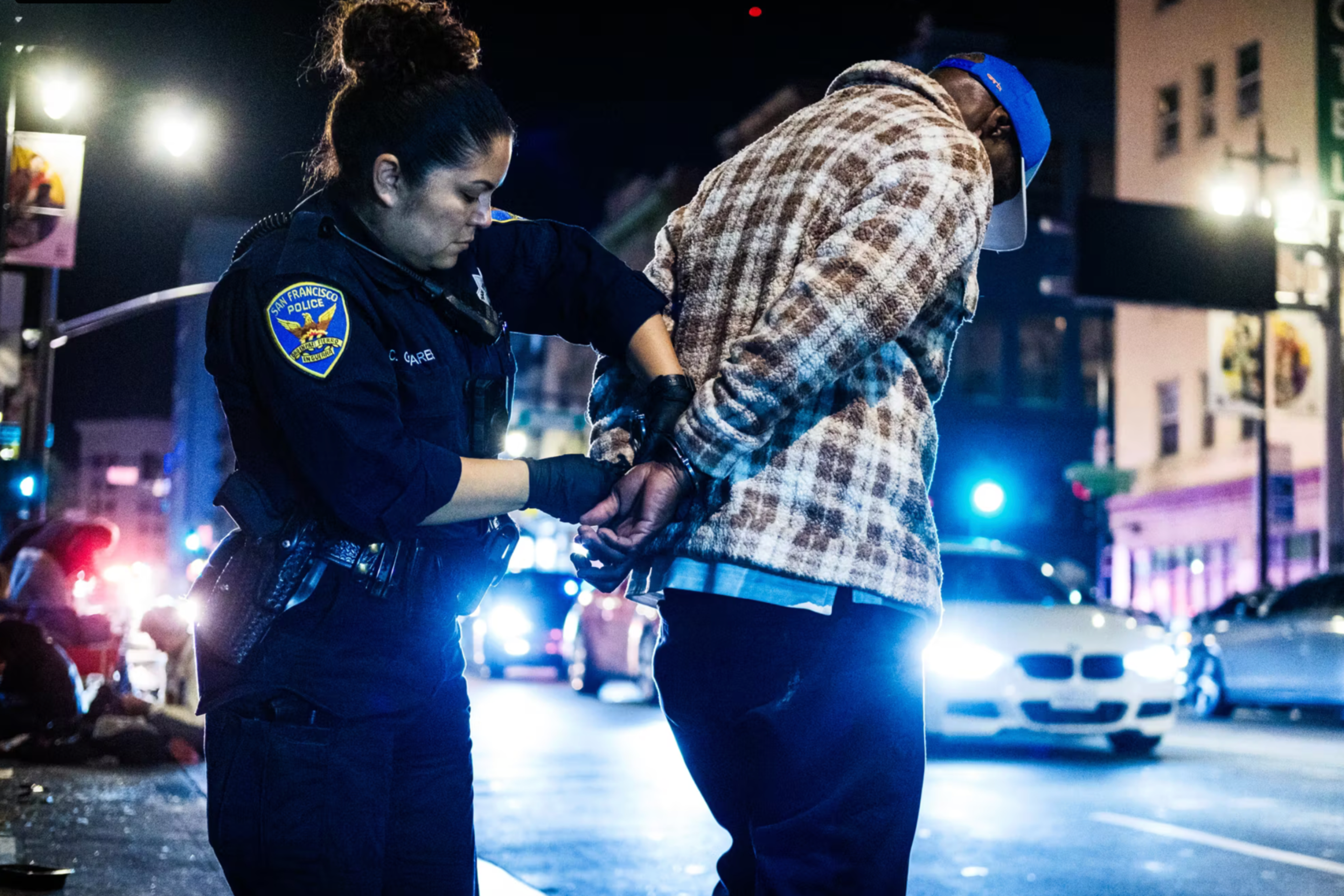 A police officer handcuffs a man wearing a plaid jacket and blue cap on a busy city street at night with cars and lights in the background.