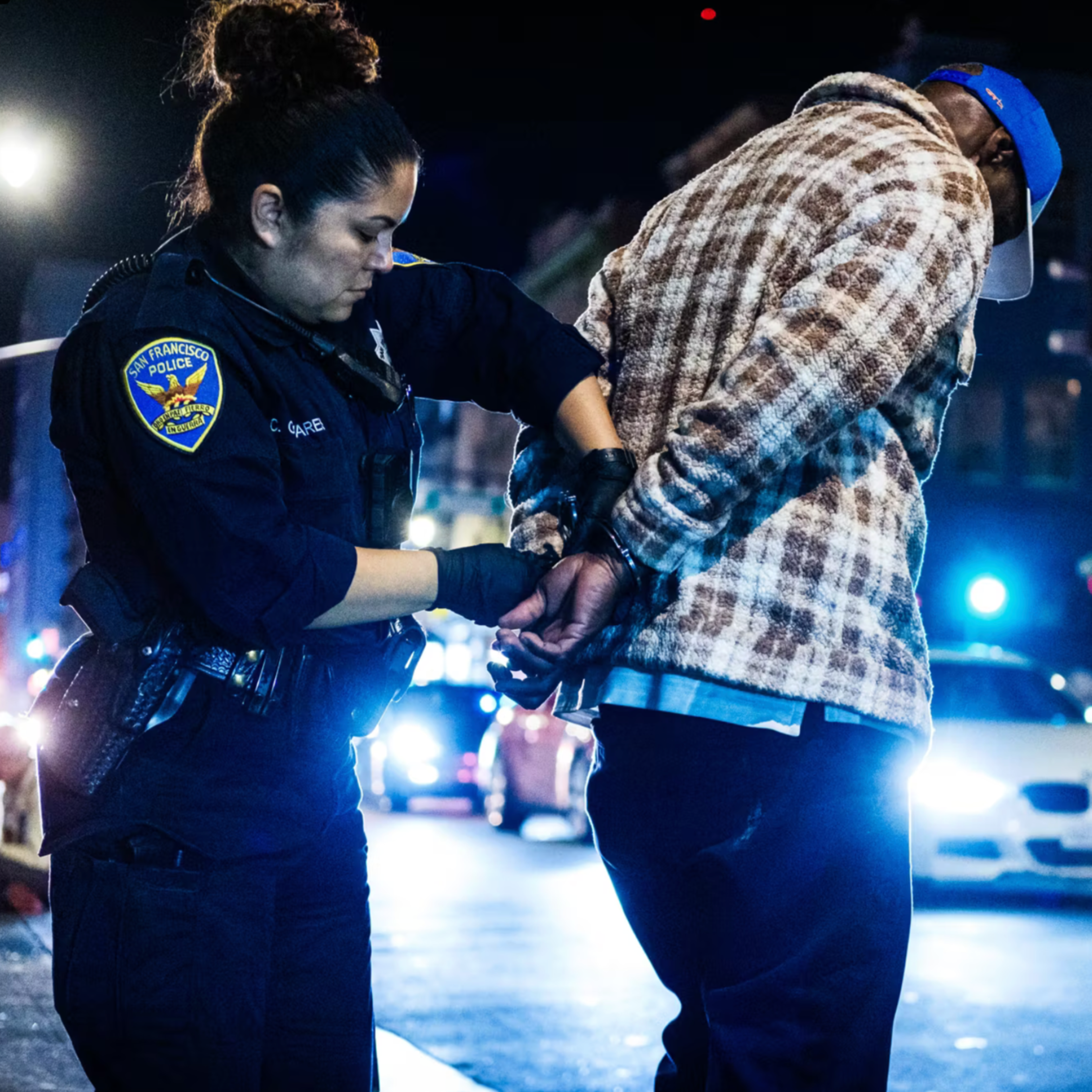 A police officer handcuffs a man wearing a plaid jacket and blue cap on a busy city street at night with cars and lights in the background.