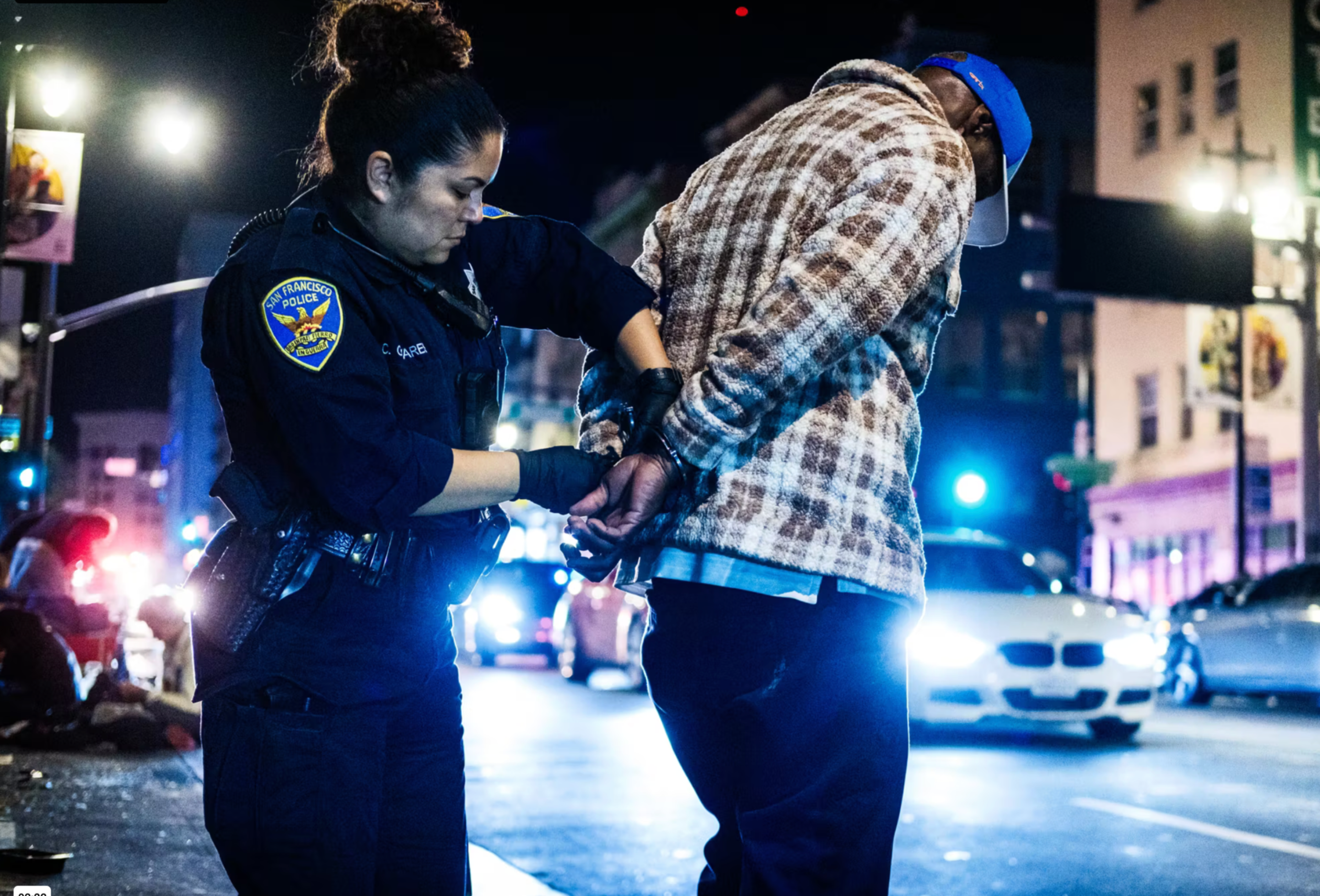 A police officer handcuffs a man wearing a plaid jacket and blue cap on a busy city street at night with cars and lights in the background.