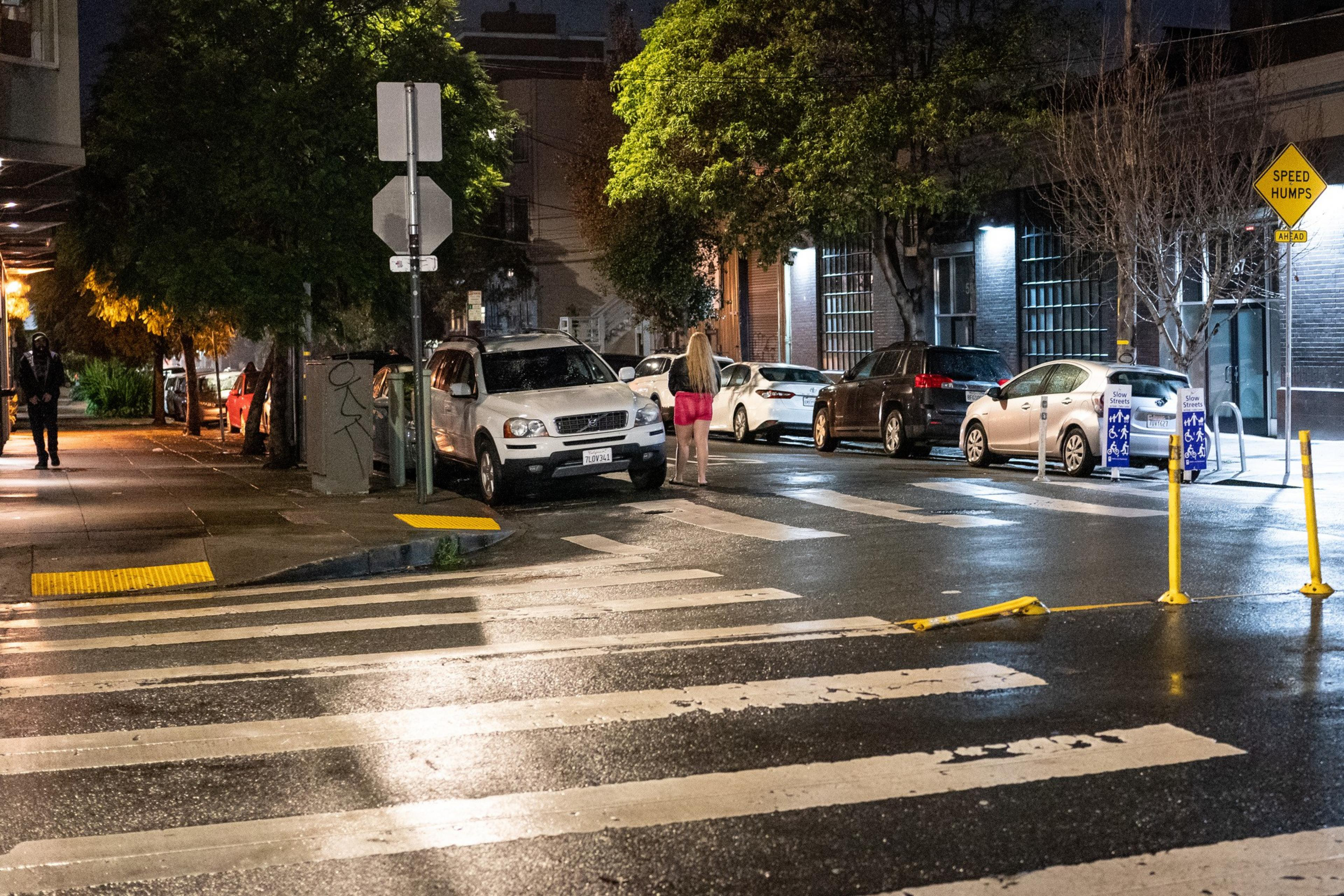 A street at night with cars, a pedestrian crossing, and a person in red shorts and a black top walking away.
